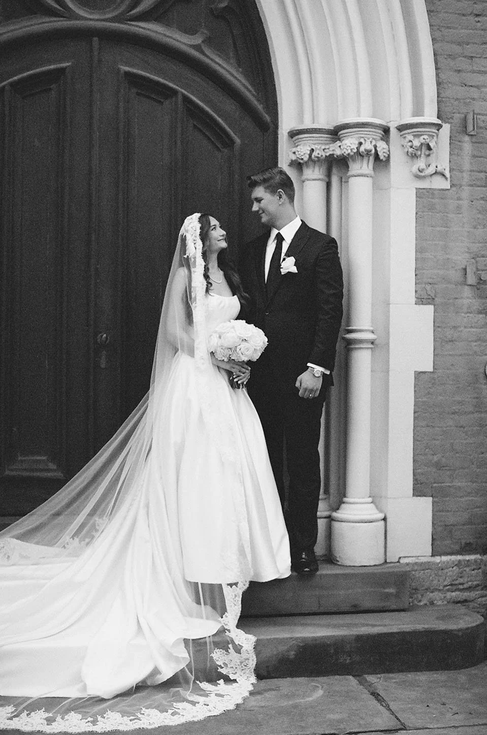 A bride and groom standing in front of a church door, gazing into each other's eyes, with the bride holding a bouquet of flowers, both dressed in wedding attire.