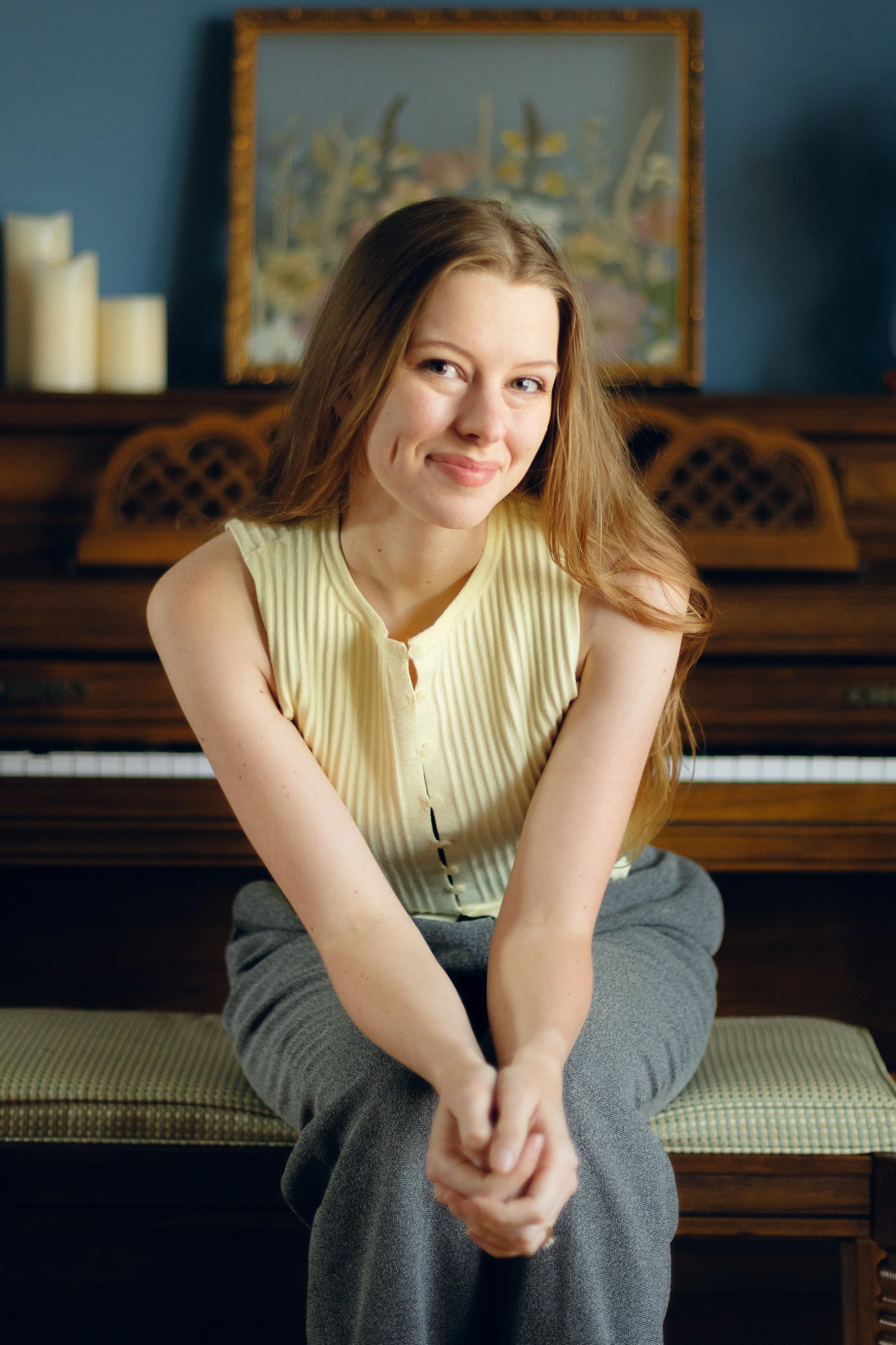 A young woman with long, light brown hair sitting cross-legged in front of a piano, smiling softly at the camera. She is wearing a sleeveless, yellowish top and gray pants. Behind her, there is a framed painting on a blue wall and white candles on the piano.