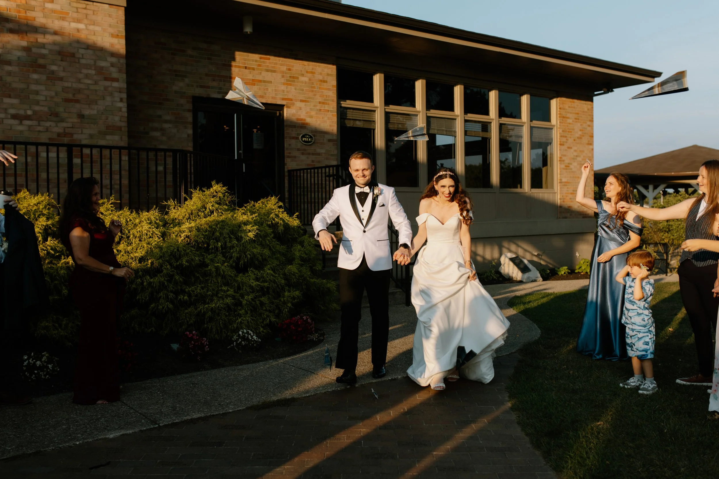 A bride and groom walking hand in hand outdoors near a brick house, surrounded by friends and family celebrating a wedding, with some guests tossing paper airplane decorations in the air during sunset.