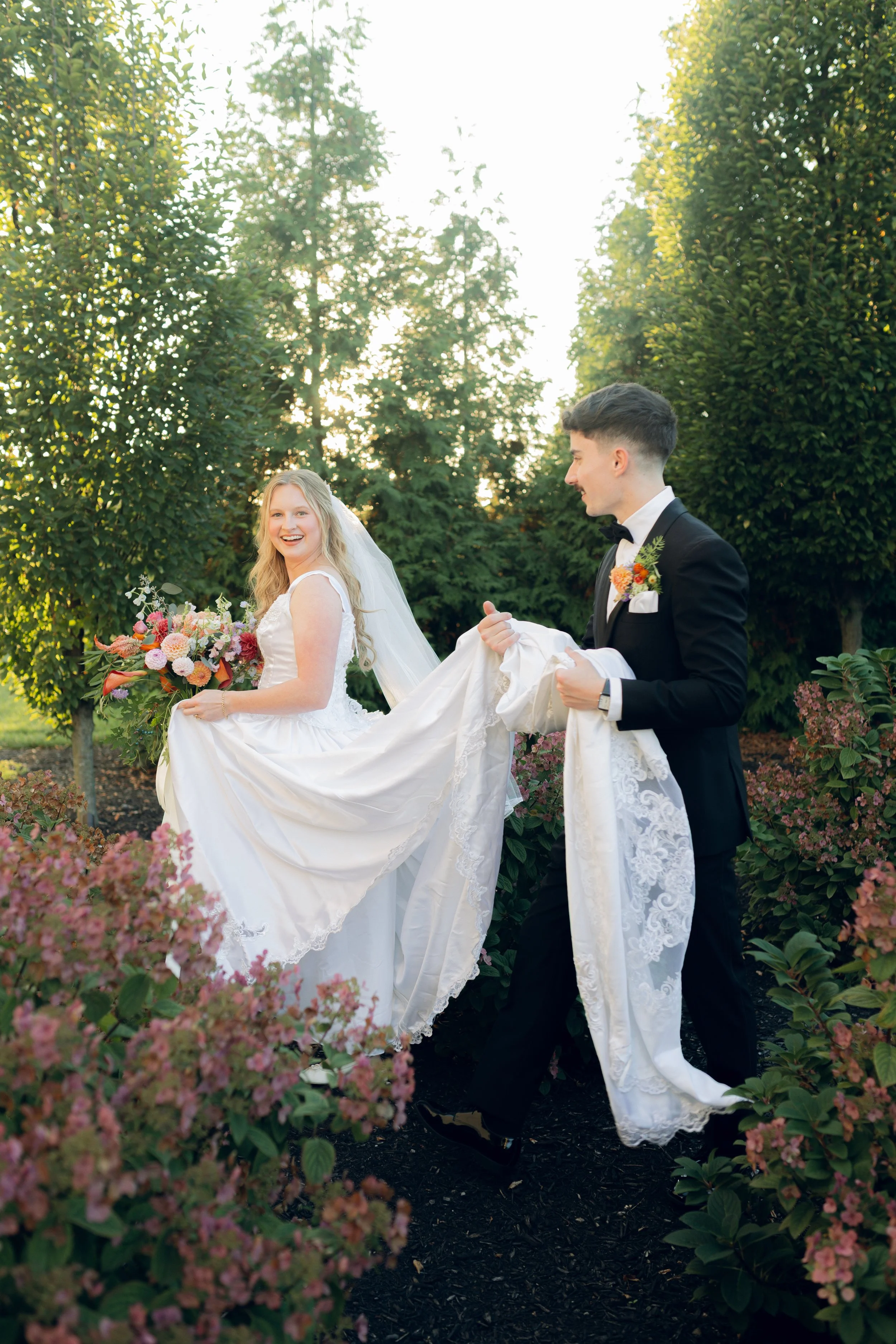 A bride and groom outdoors, with the groom lifting the bride's wedding dress train. The bride is holding a bouquet, and both are smiling. The scene is surrounded by greenery and pink flowers, with sunlight filtering through trees.