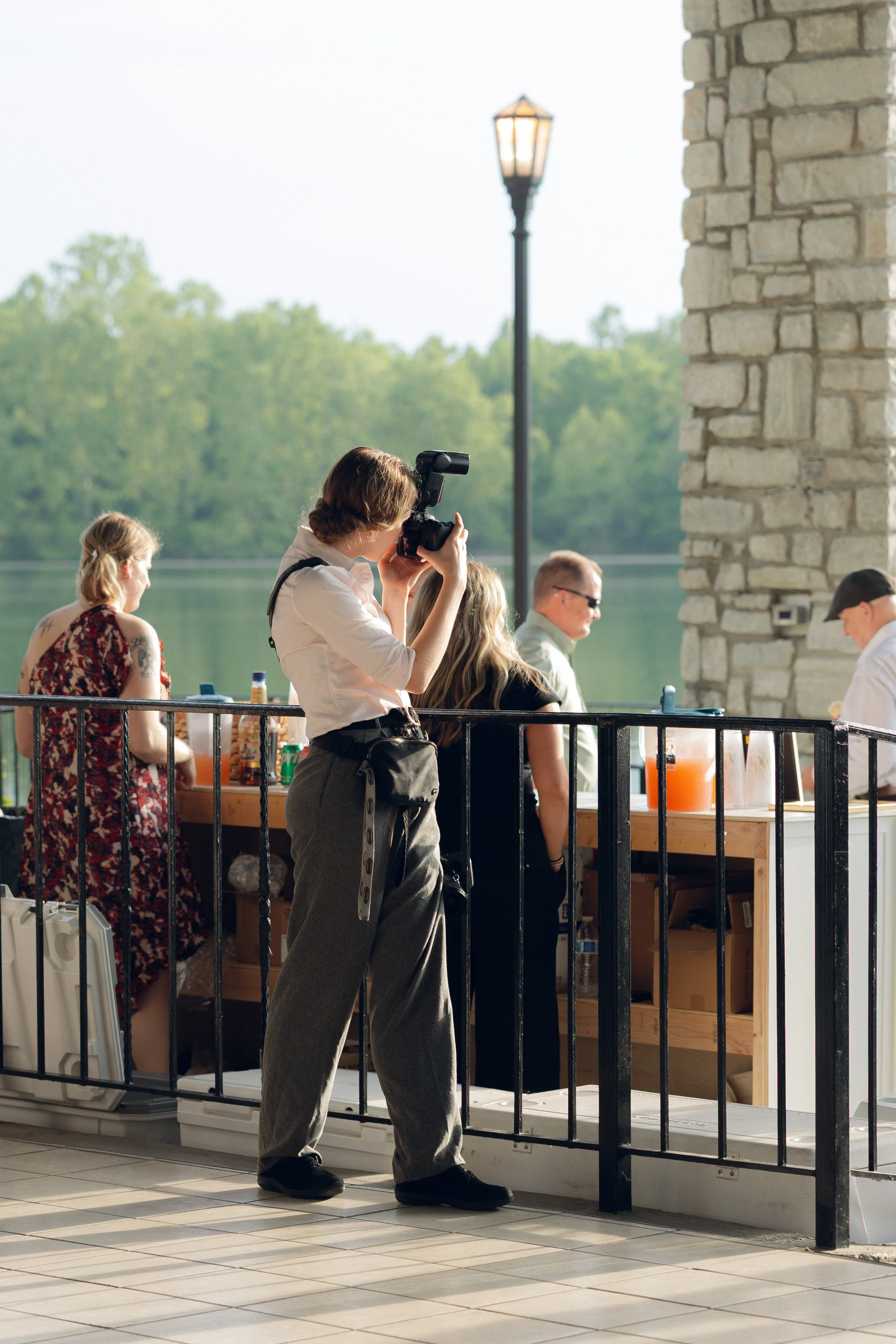 A woman with shoulder-length brown hair, wearing a light pink shirt, gray pants, and black shoes, is taking a photo with a camera. She stands near a black metal railing outside near a stone wall and a table with drinks, with three people sitting at the table. There is a lamppost and trees in the background.
