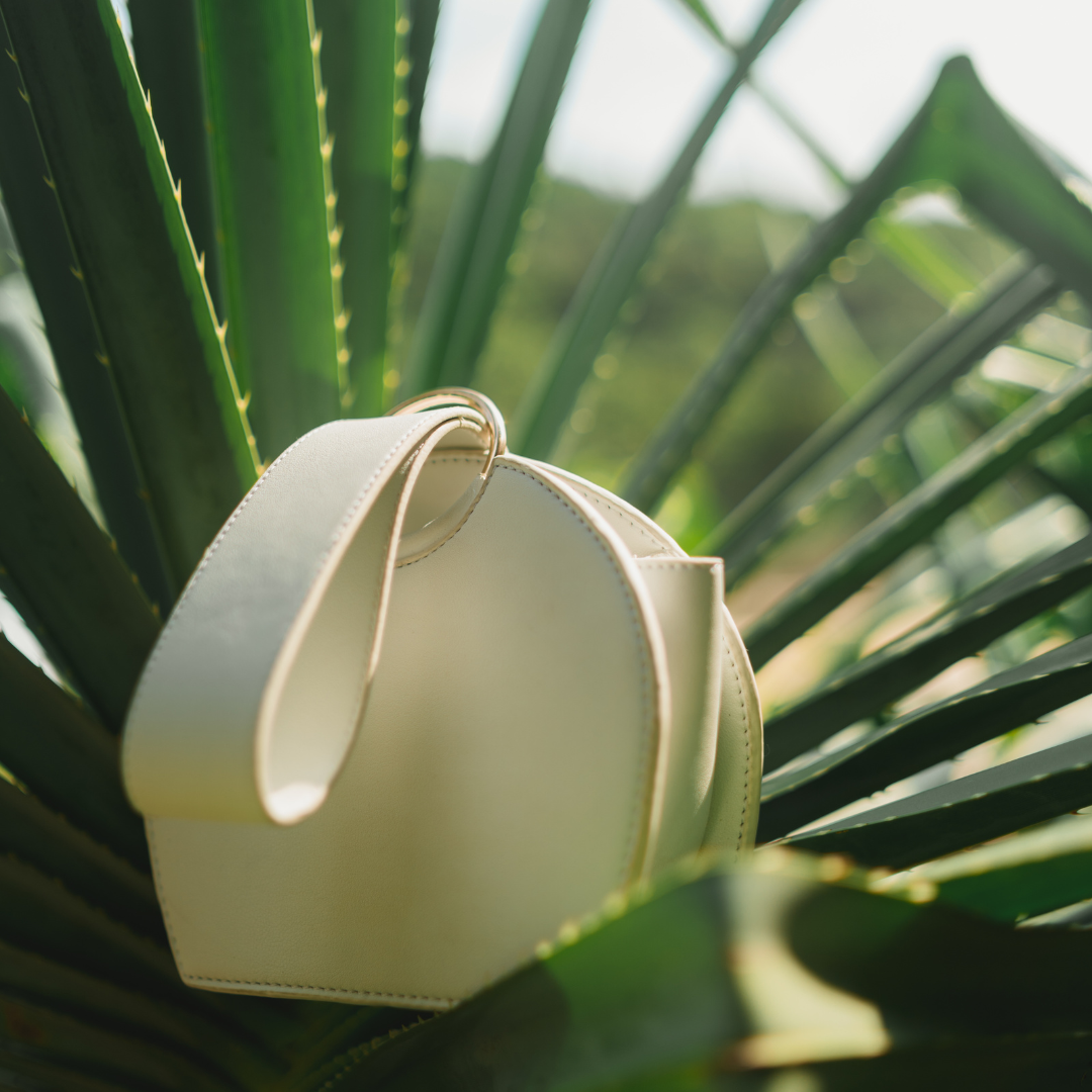 A cream-colored handbag hanging on green plant leaves outdoors.