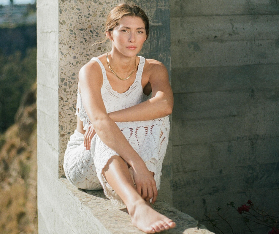 A young woman with light brown hair, wearing a white crocheted dress and a gold necklace, sitting barefoot on a concrete ledge outdoors with a neutral expression.