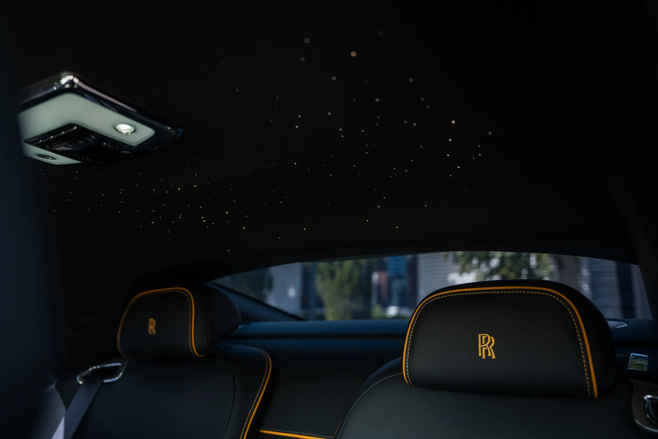 The interior of a luxury car showing a starry night sky ceiling, black leather headrests with yellow stitching and Rolls Royce initials, and a rearview mirror.