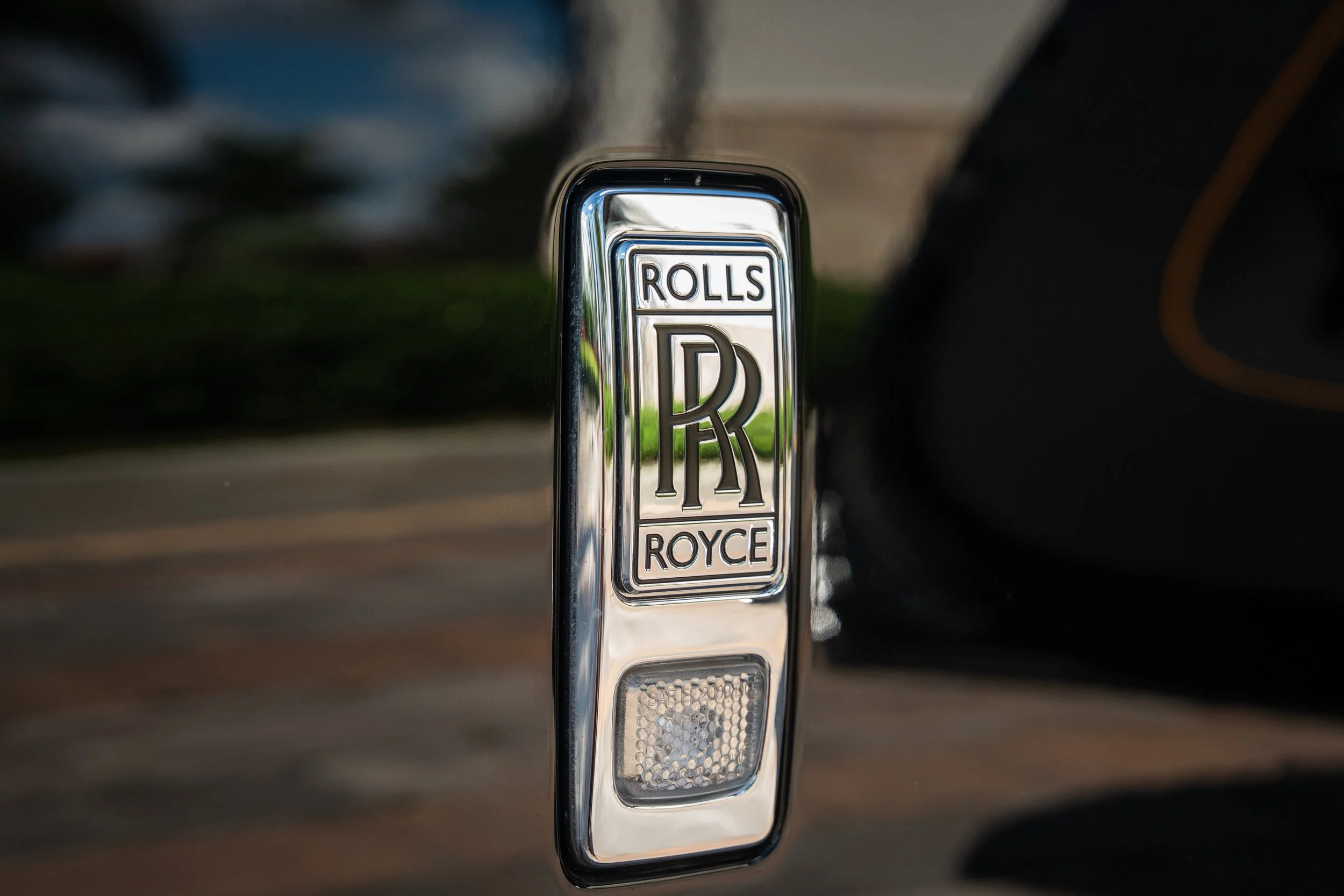 Close-up of a shiny silver Rolls-Royce emblem on a car, displaying the brand's logo with the words 'ROLLS' and 'ROYCE' above and below a large 'RR' monogram.