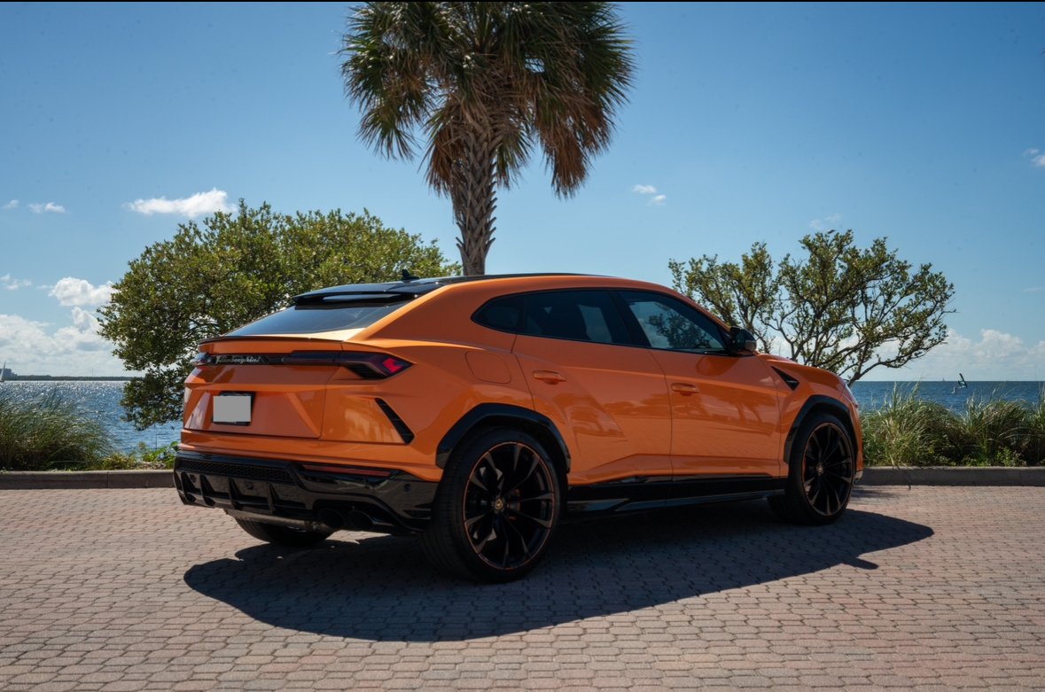 An orange luxury SUV parked on a brick road near a body of water, with palm trees and greenery in the background under a clear blue sky.