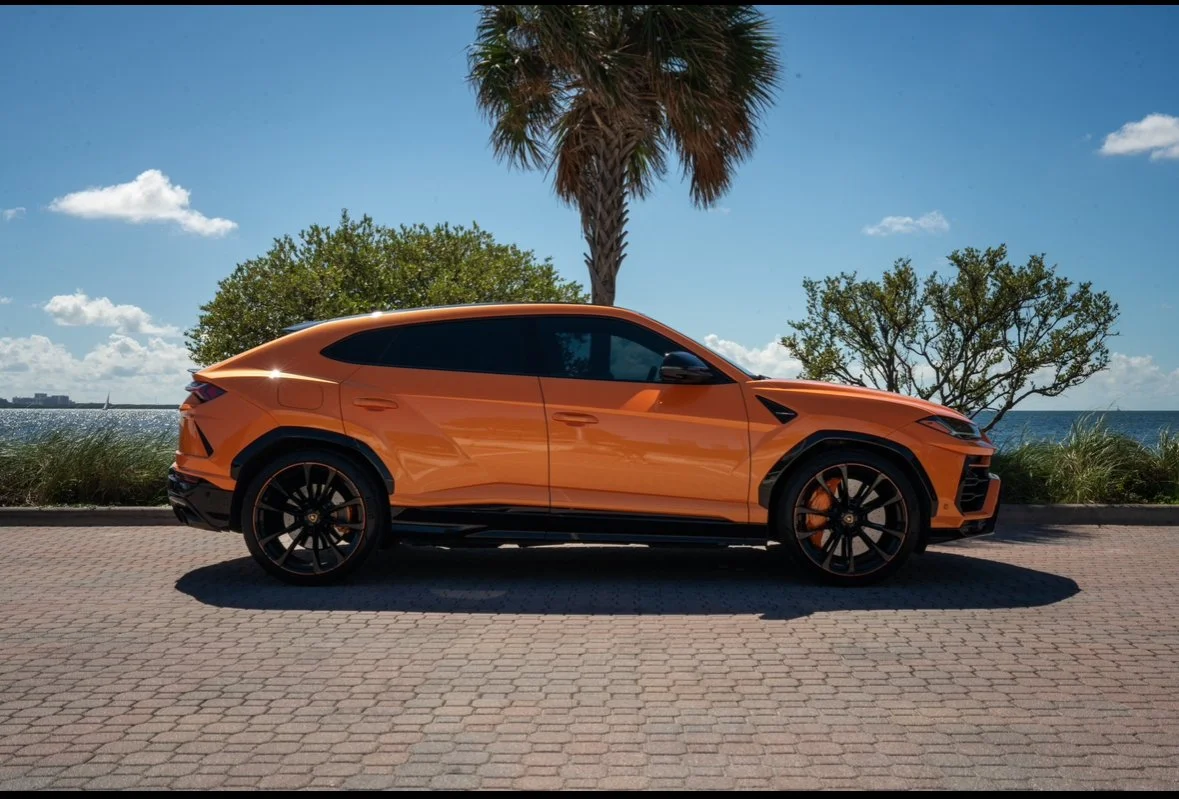 An orange luxury SUV parked on a paved road near the beach, with palm trees and a blue sky in the background.