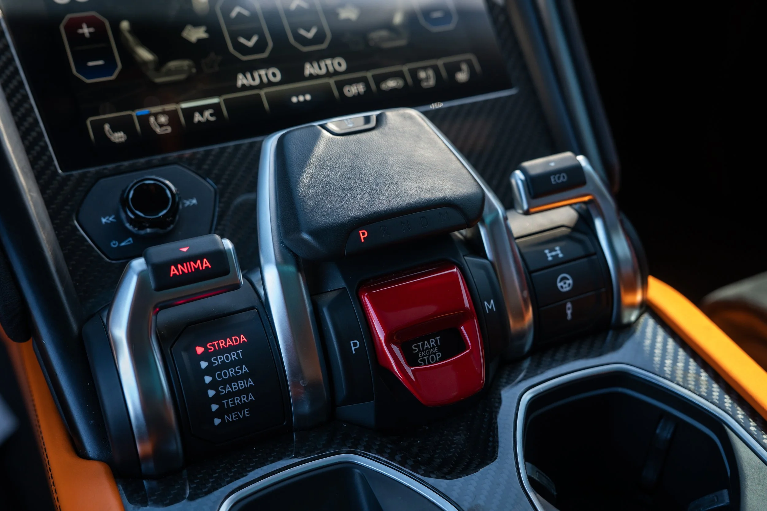 Close-up of a luxury car's center console with various buttons, switches, and an electronic screen, including a red start/stop button, gear selector, driving mode selector, and climate controls, with a carbon fiber finish.