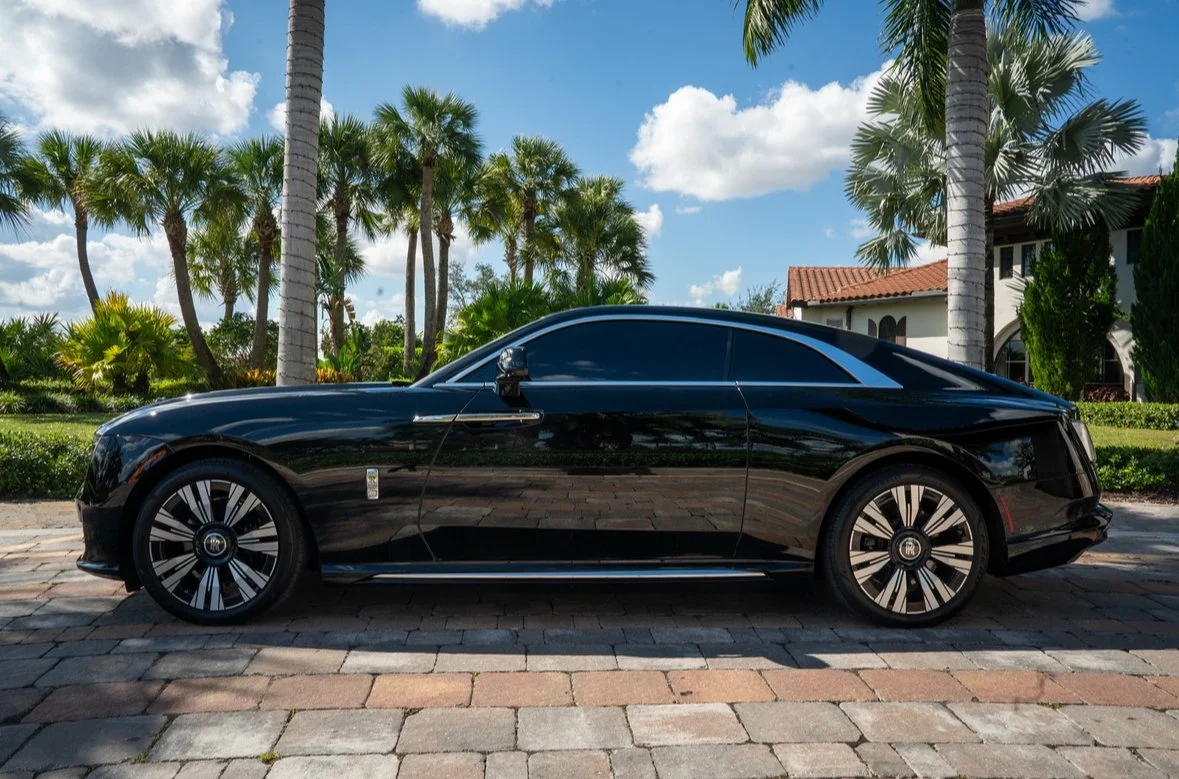 Black luxury coupe car parked on cobblestone street with palm trees and a house in the background.