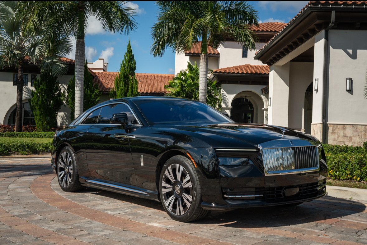 Black luxury car parked on a cobblestone driveway in front of a modern house with palm trees and blue sky.