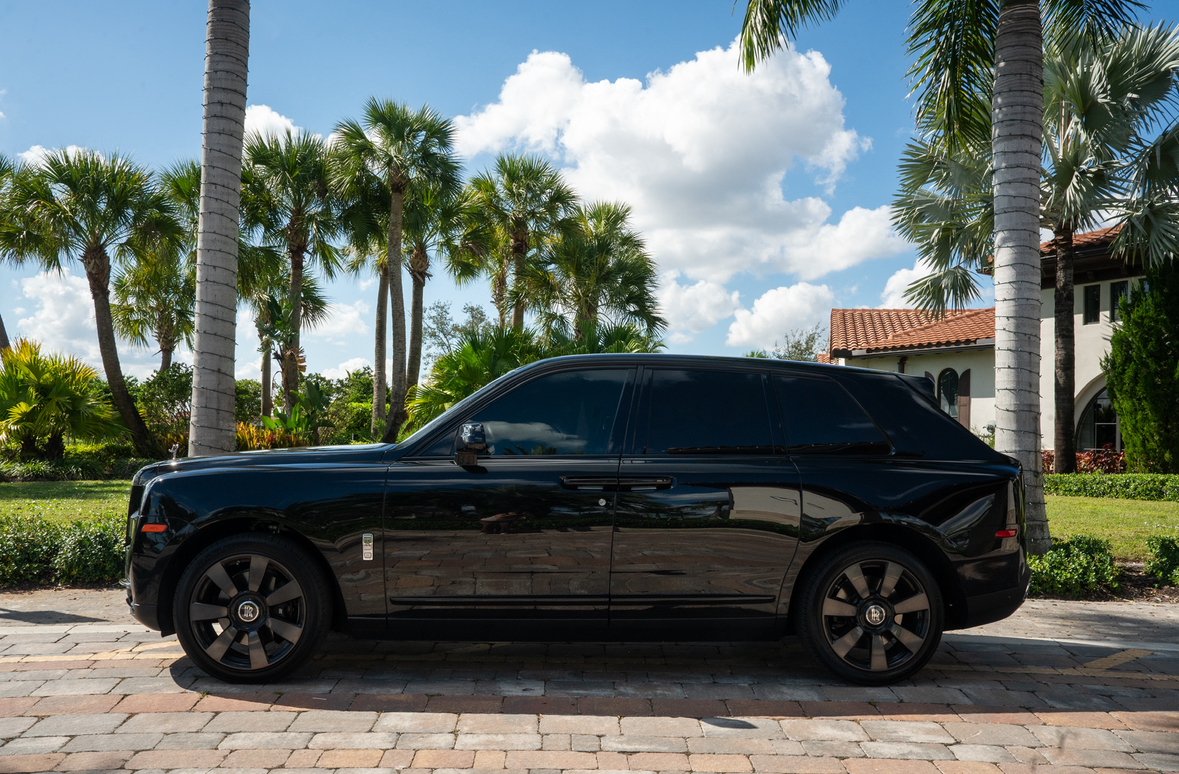 A black luxury SUV parked on a brick driveway in front of a tropical landscape with palm trees and a white house with a red-tiled roof under a partly cloudy sky.