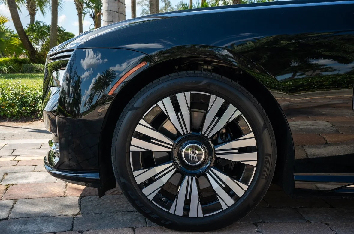Close-up of a black luxury car's front wheel and fender, with reflective surface showing trees and sky outside.