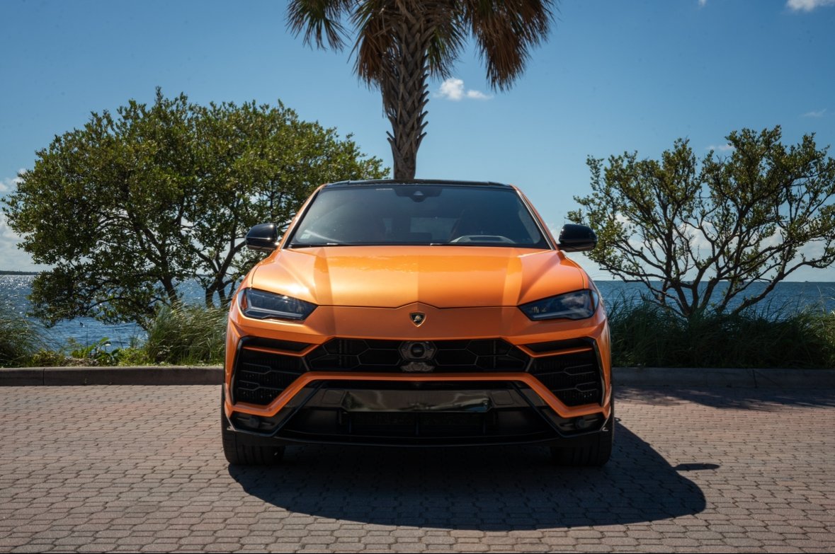 Front view of an orange Lamborghini Urus parked on a brick paved area near the water, with trees, a palm tree, and the ocean in the background.
