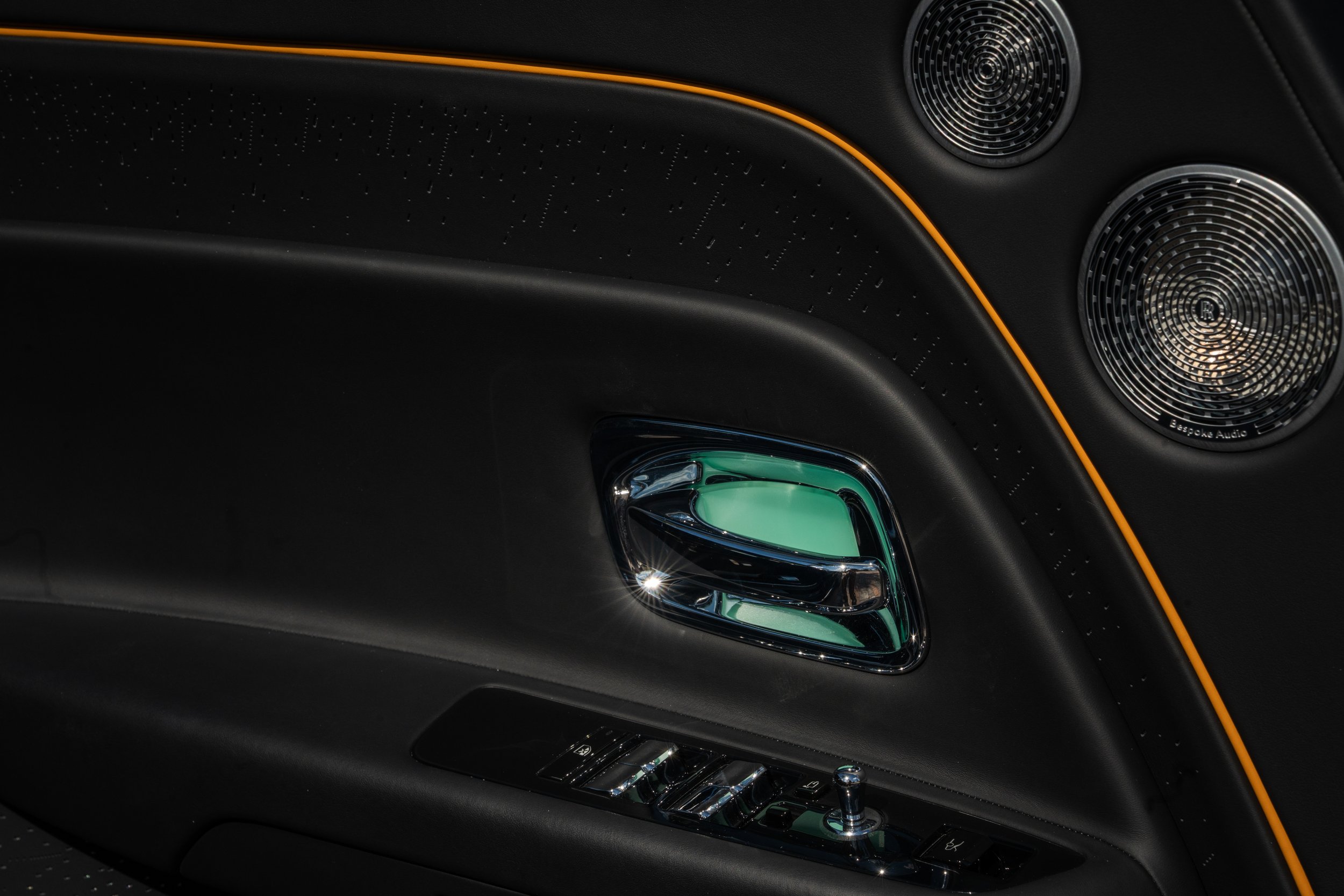 Close-up of an interior car door panel featuring a metallic door handle, window controls, and a speaker, with accents of orange stitching.