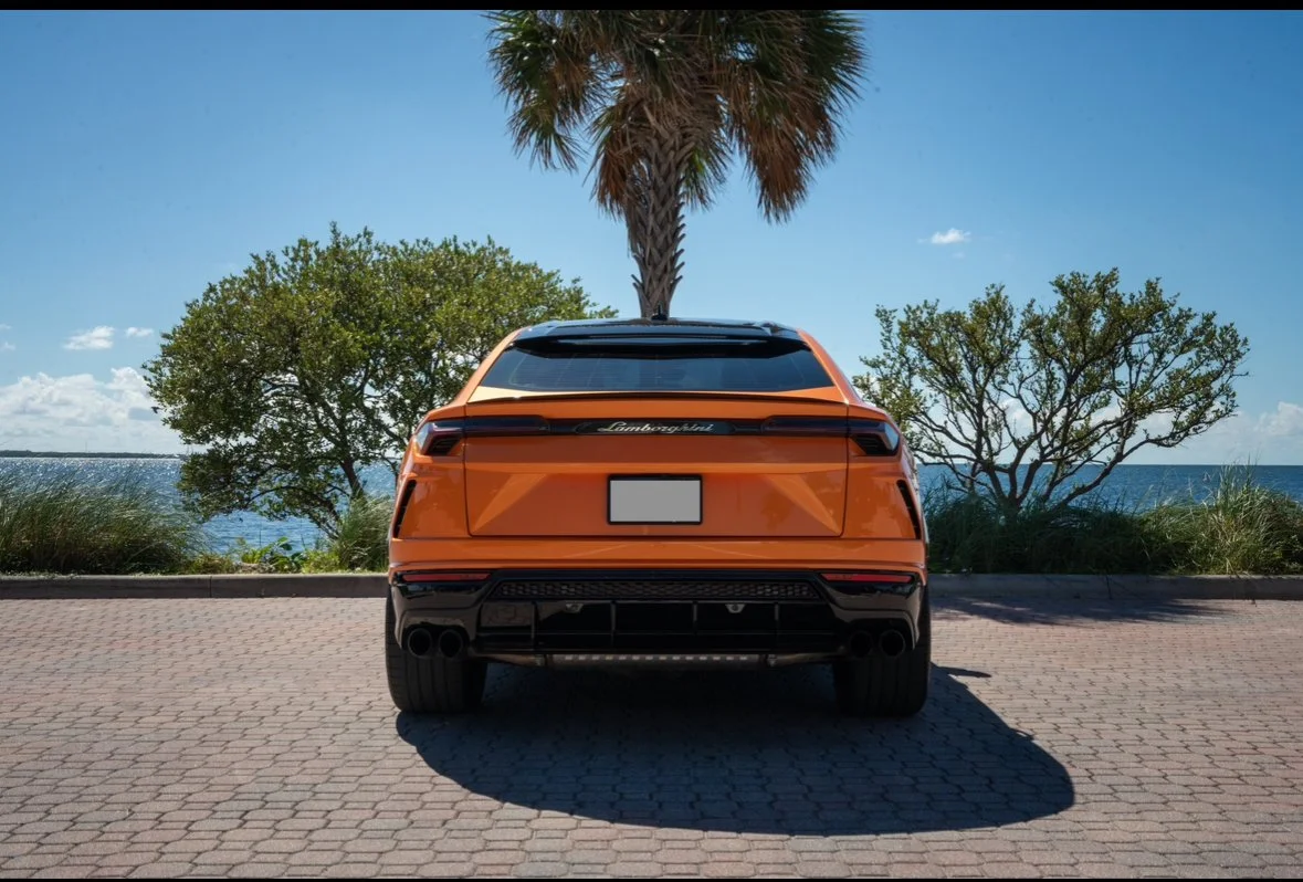 Orange Lamborghini SUV parked on a brick promenade near the water, with trees and a palm tree behind it and a clear blue sky.