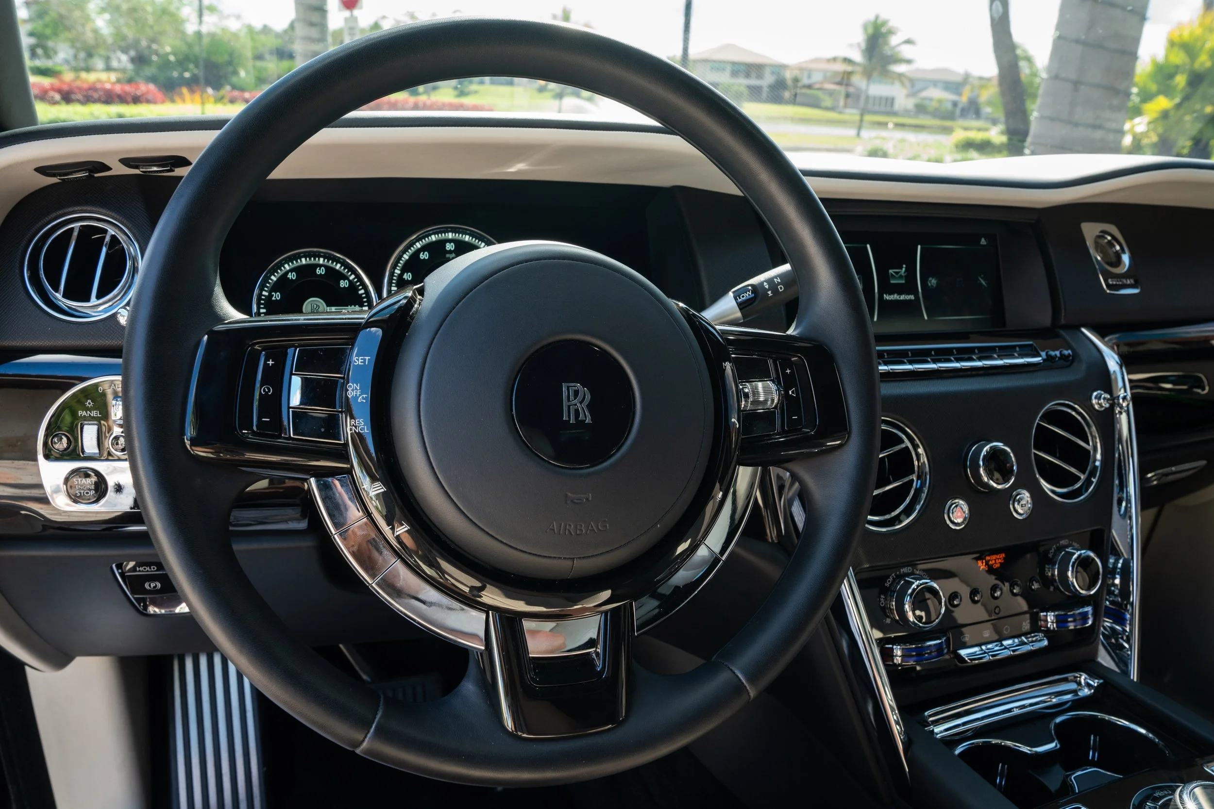 Interior of a luxury car, showing a steering wheel with a Rolls-Royce logo, dashboard with instrument cluster, air vents, and control panel, with sunlight streaming through the windows.