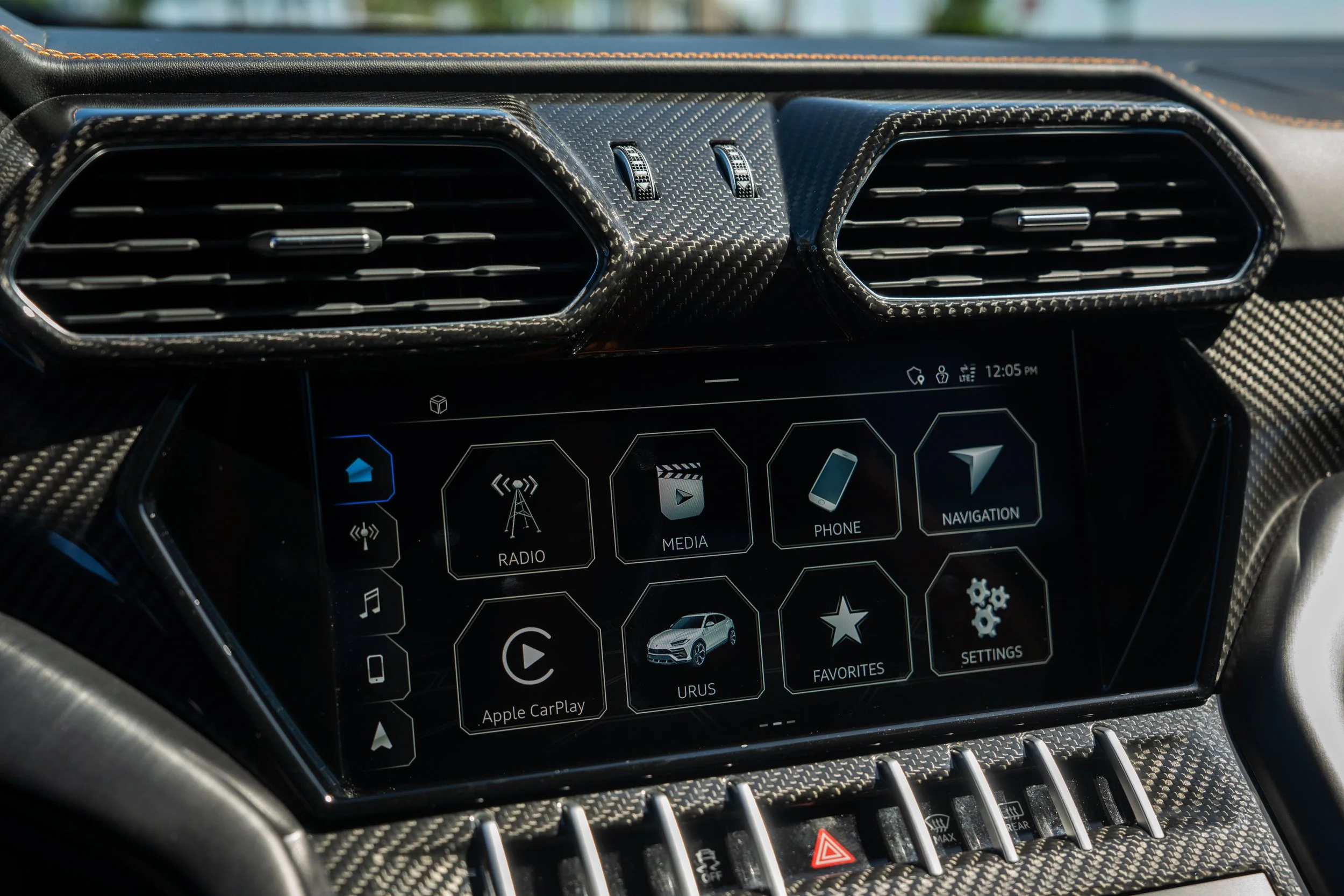 Car dashboard display with touchscreen interface showing navigation, radio, media, phone, favorites, settings, and Apple CarPlay options; surrounding vents with carbon fiber trim.