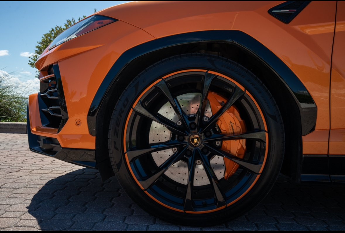 Close-up of an orange Lamborghini sports car's front wheel with black accents and a perforated brake disc visible through the rim.