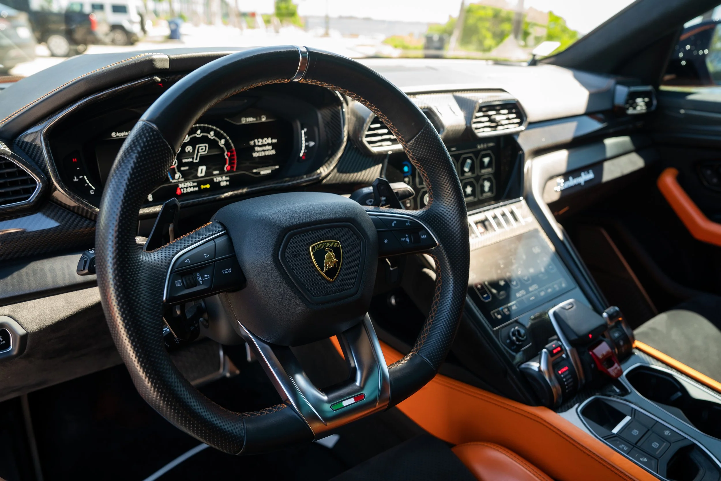 Interior of a Lamborghini sports car, featuring a steering wheel with the Lamborghini logo, digital dashboard, and control panel with orange accents.