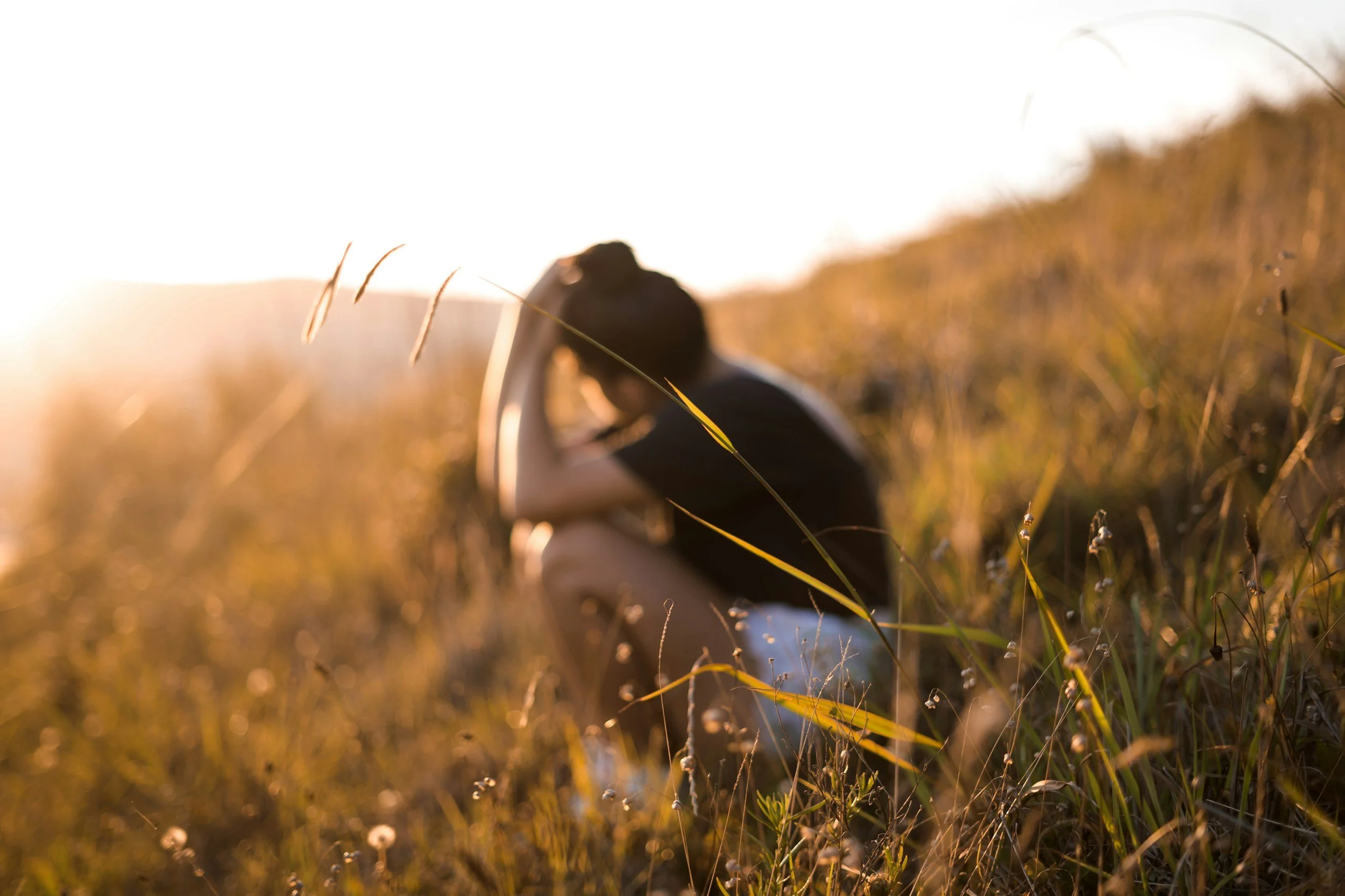 Young adult, with dark hair, crouching down in an open field, with hands on her head, representing a young adult struggling with the pains of estrangement and may benefit from online and in-person mental health therapy for estrangement