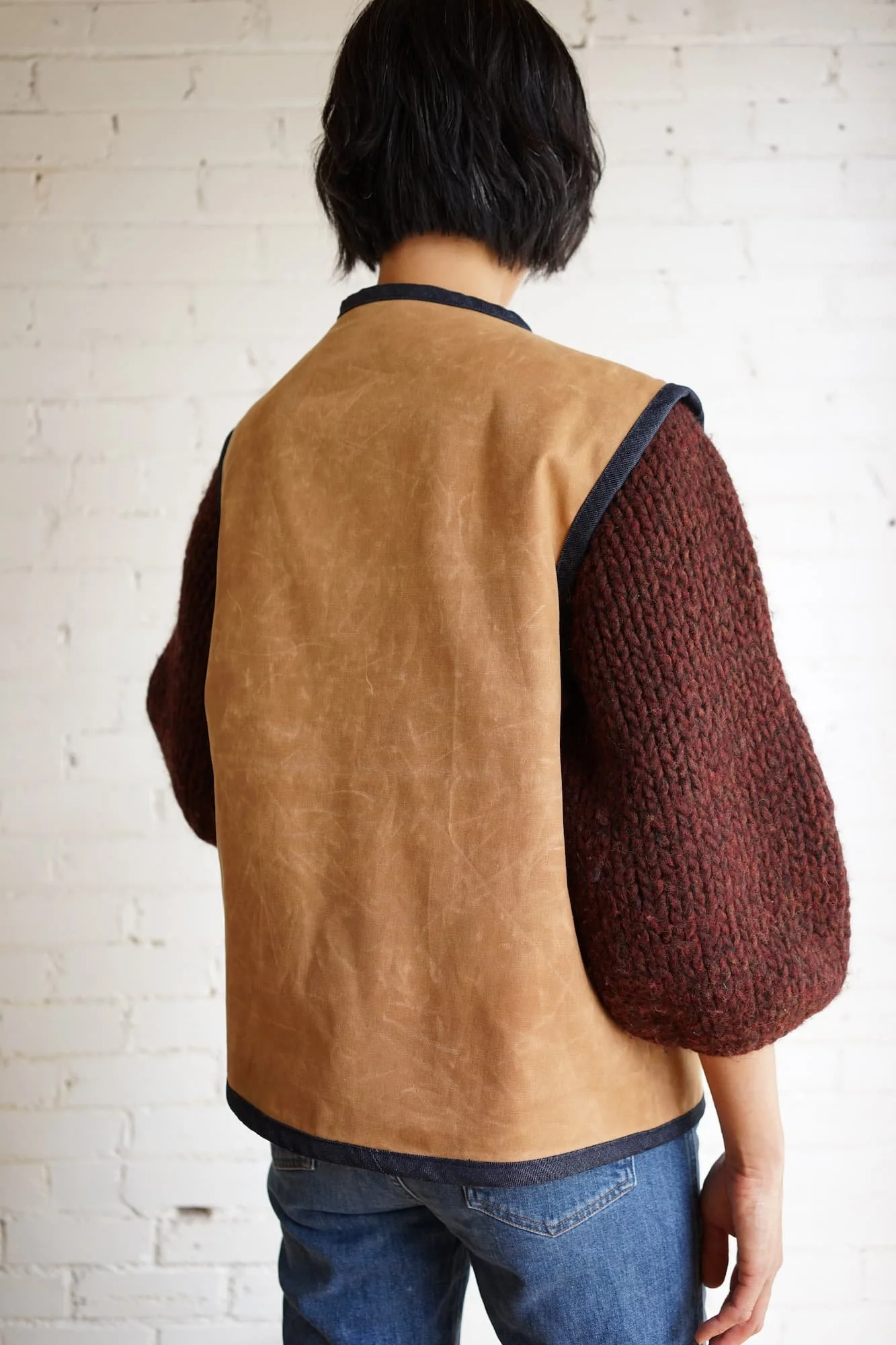Back view of tan waxed canvas Nomad Utility Vest with navy denim binding, worn over a burgundy knit sweater