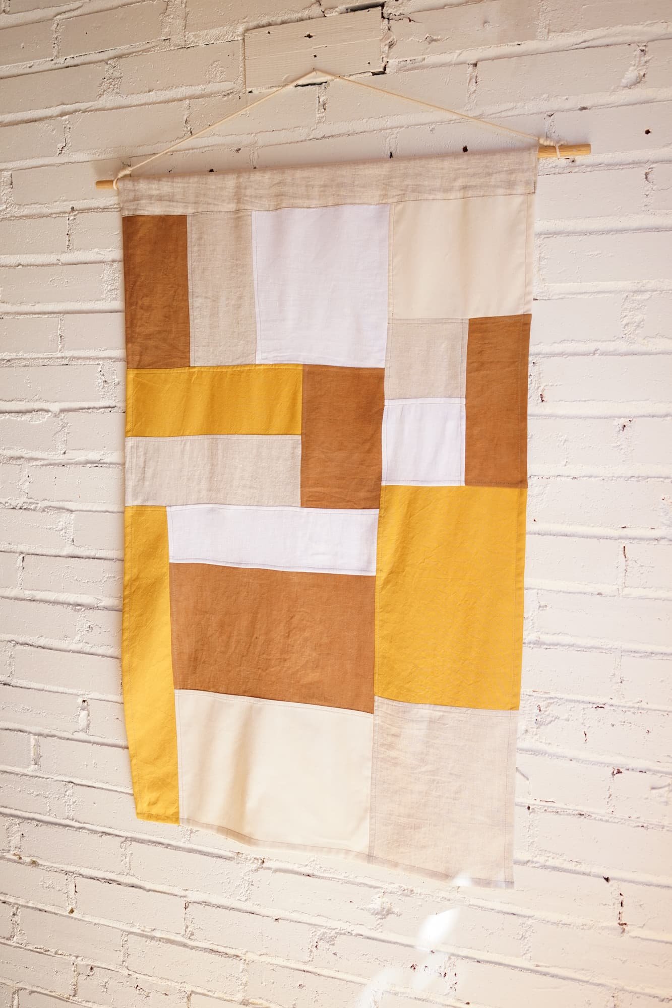 Amber and mustard jogakbo linen wall hanging with cream and white sections, displayed on a wooden dowel against a whitewashed brick wall