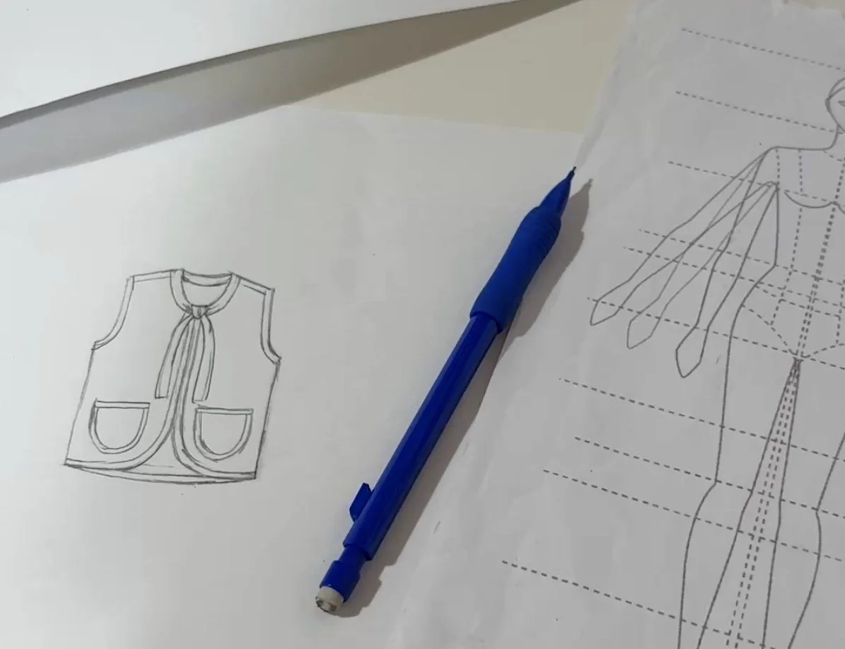 Early pencil sketch of the Nomad Utility Vest showing a V-neck vest with bound edges, a tie at the neckline, and two rounded patch pockets, next to a blue mechanical pencil and a fashion figure template