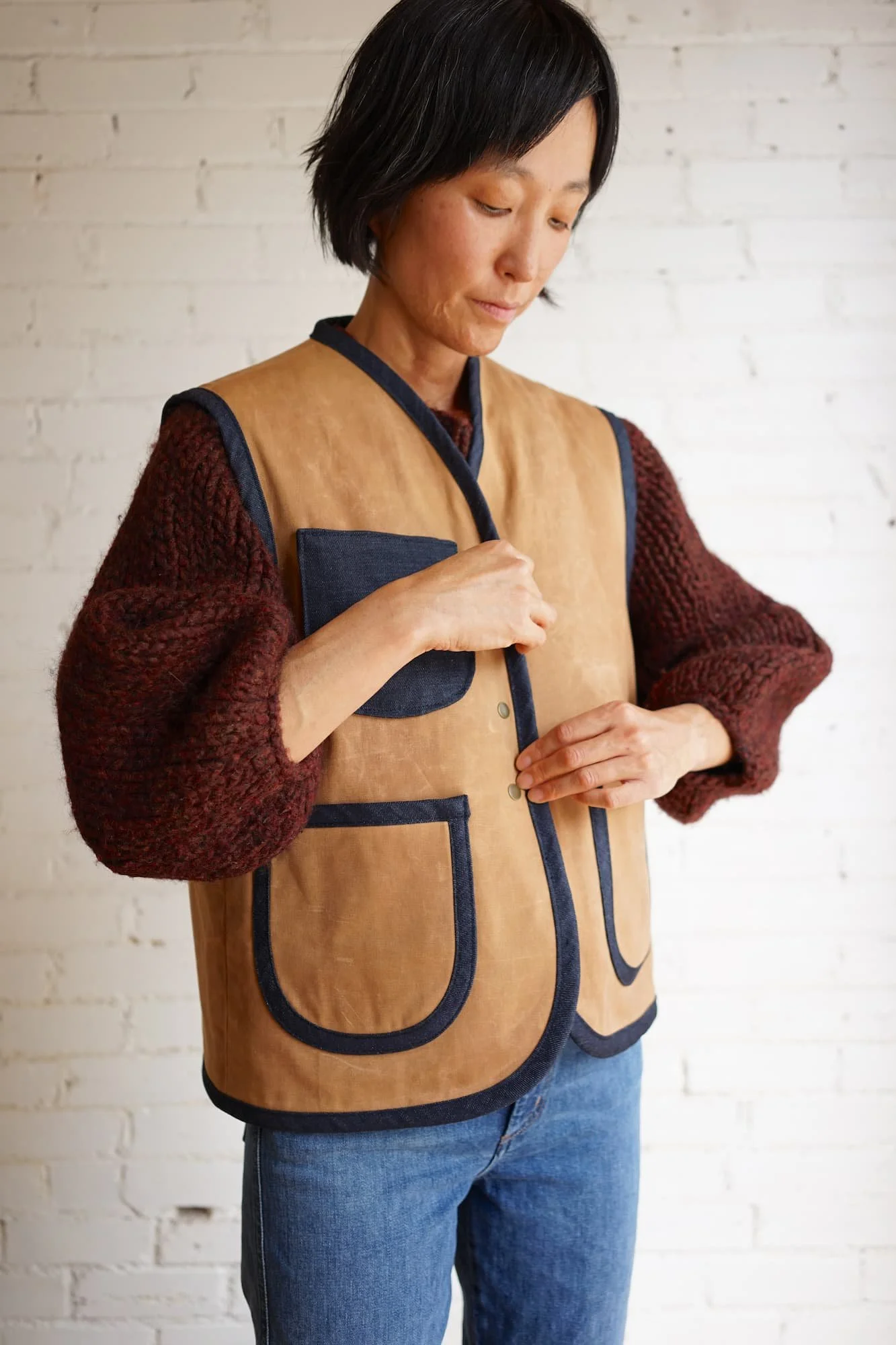Woman fastening brass snap closures on tan waxed canvas Nomad Utility Vest with navy denim binding and pockets