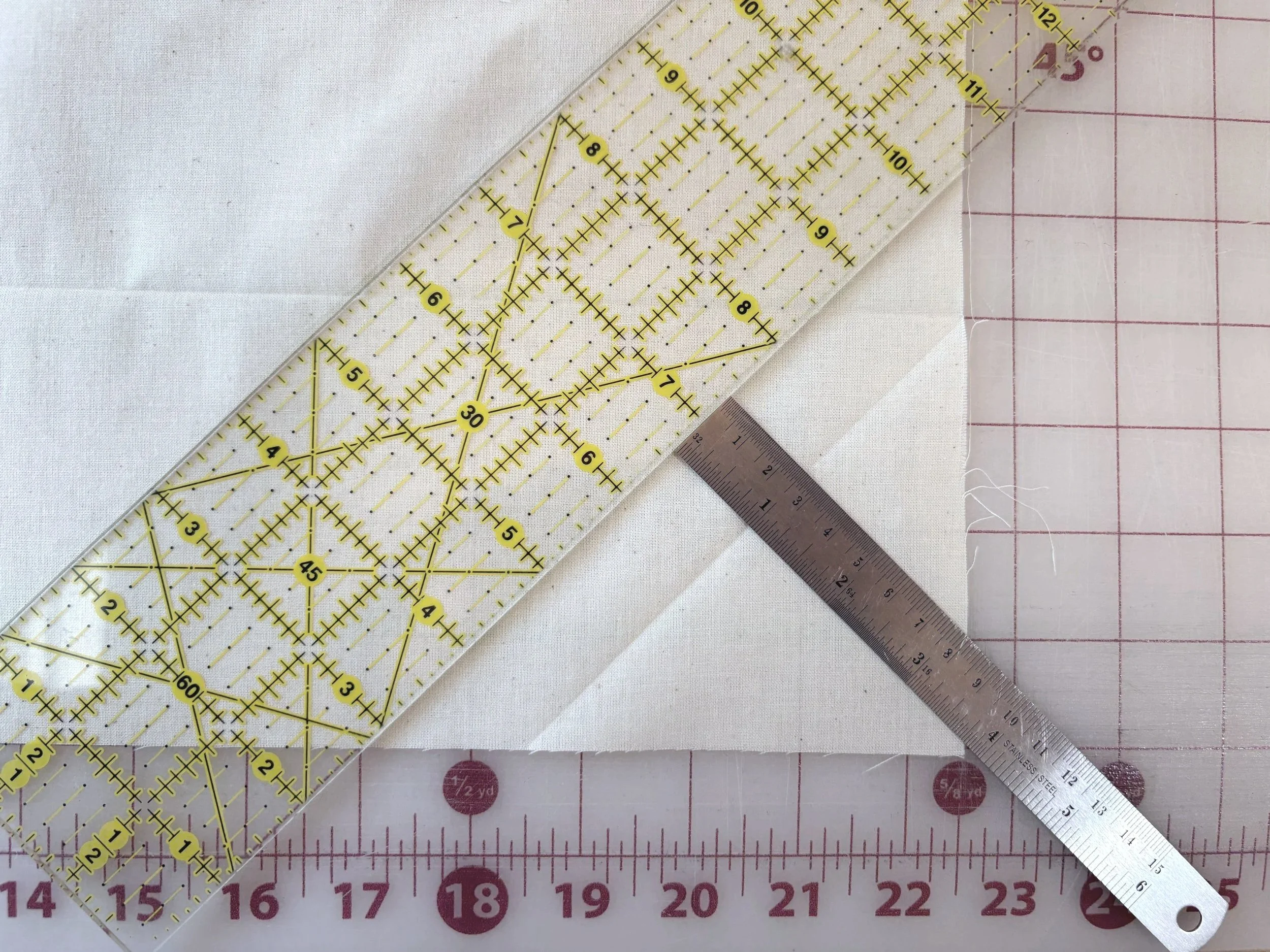 Quilting ruler with 45-degree gridlines placed over the diagonal fabric fold to verify the true bias angle, with a metal ruler positioned to measure strip width