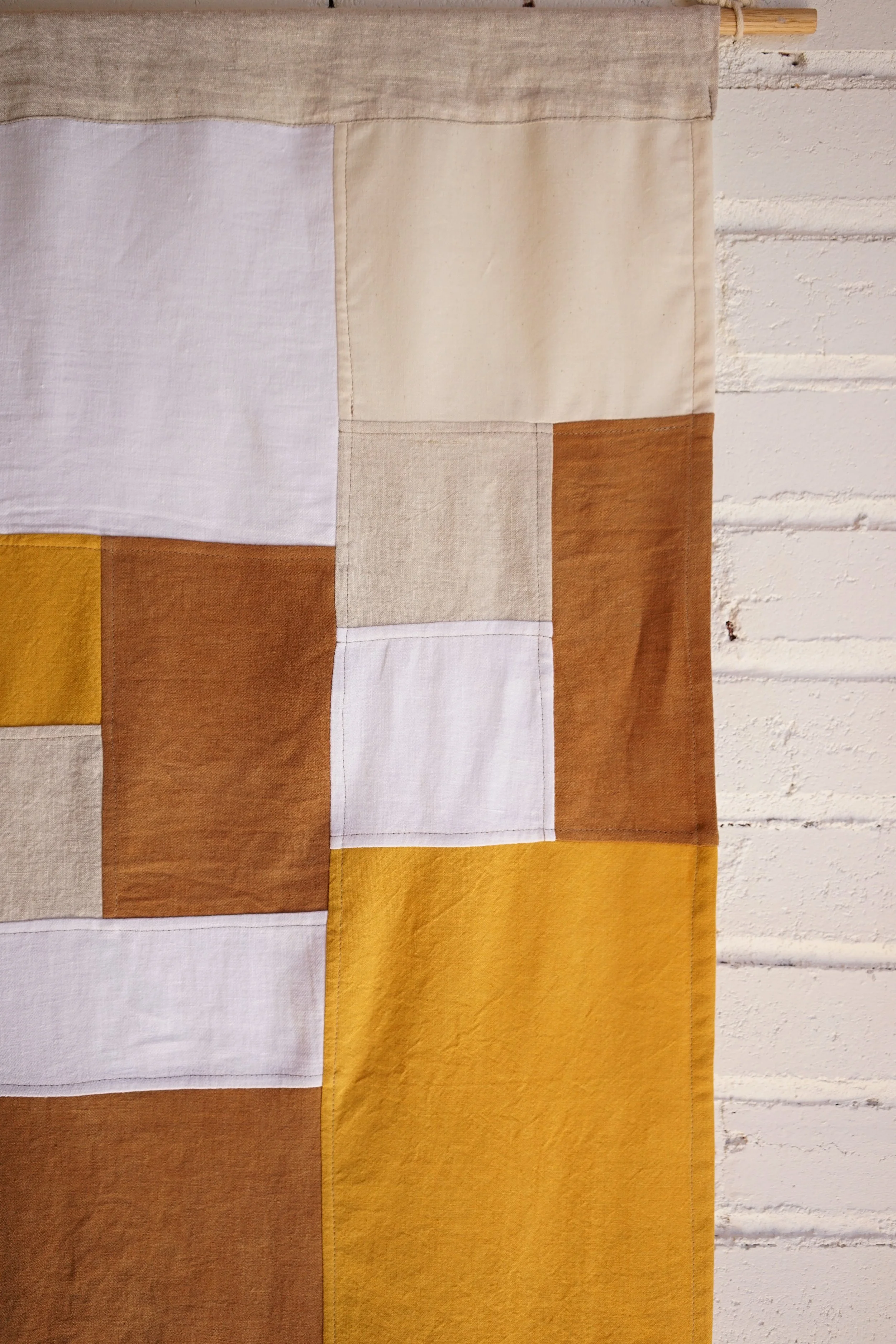 Detail of amber jogakbo linen wall hanging near wooden hanging dowel against painted brick wall