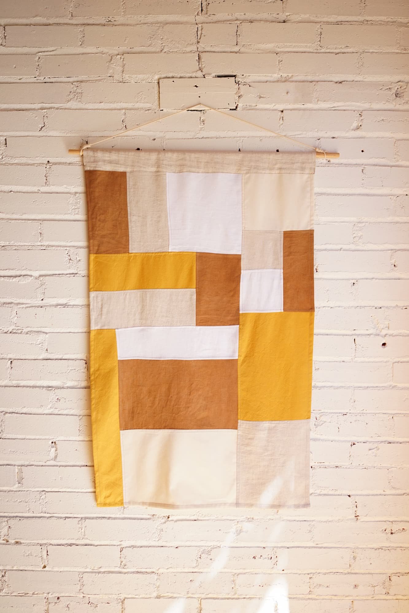 Amber and mustard jogakbo linen wall hanging, photographed straight-on, with cream and white sections displayed on a wooden dowel against a whitewashed brick wall
