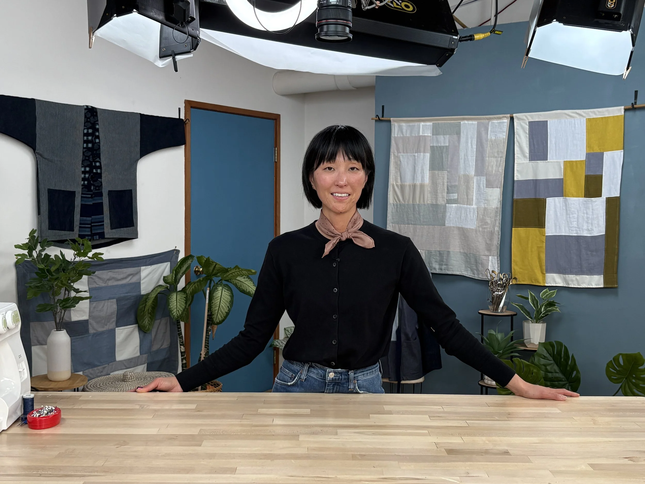 Thanks for the correction — updated:  `Mimi Shim standing behind a wooden work table at a CreativeBug production studio, with a patchwork jacket and two patchwork textile pieces hanging on the walls behind her, under studio lighting