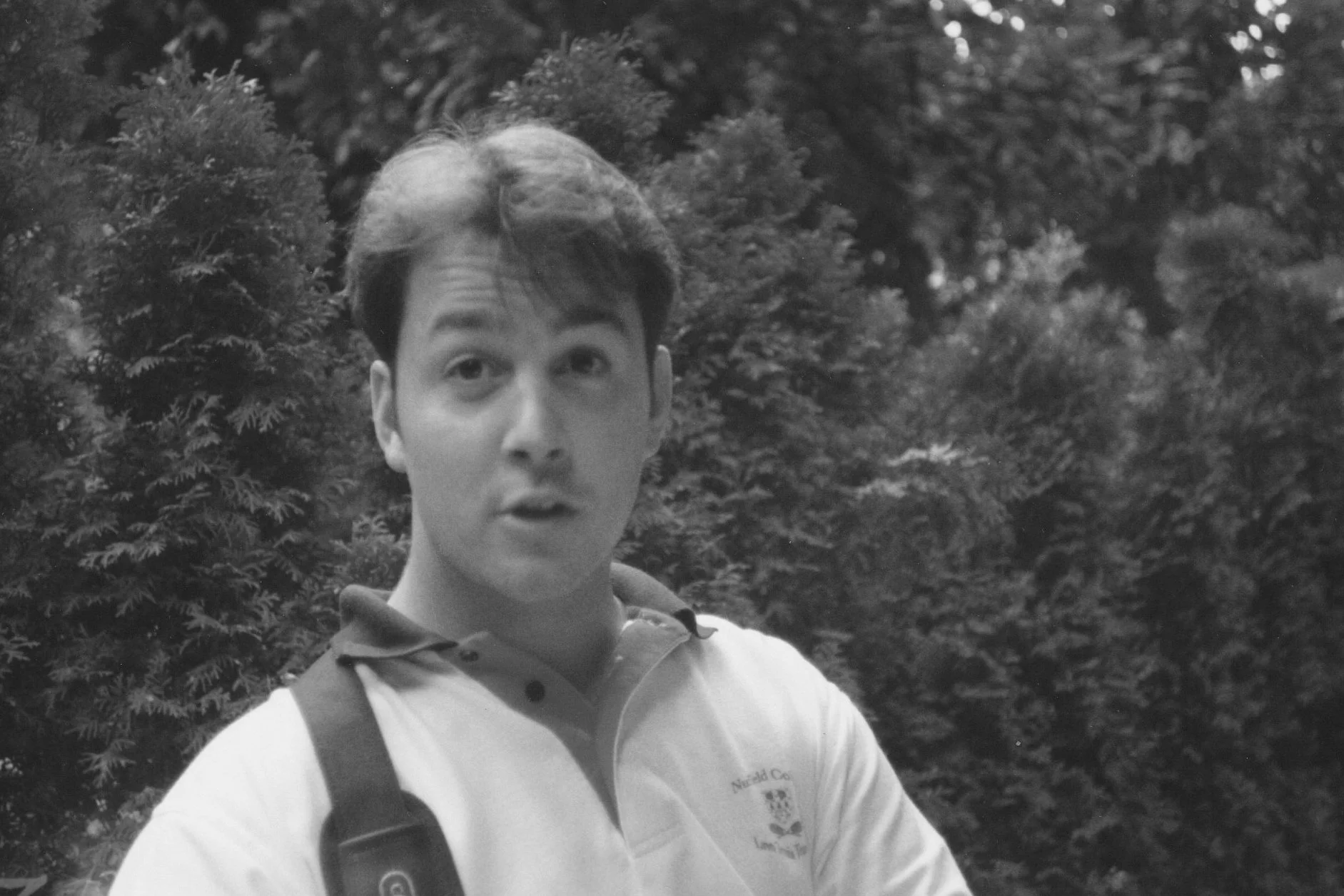 A young man with dark, slightly wavy hair and fair skin standing outdoors in front of trees and bushes, wearing a polo shirt with a logo and carrying a bag strap over his shoulder.