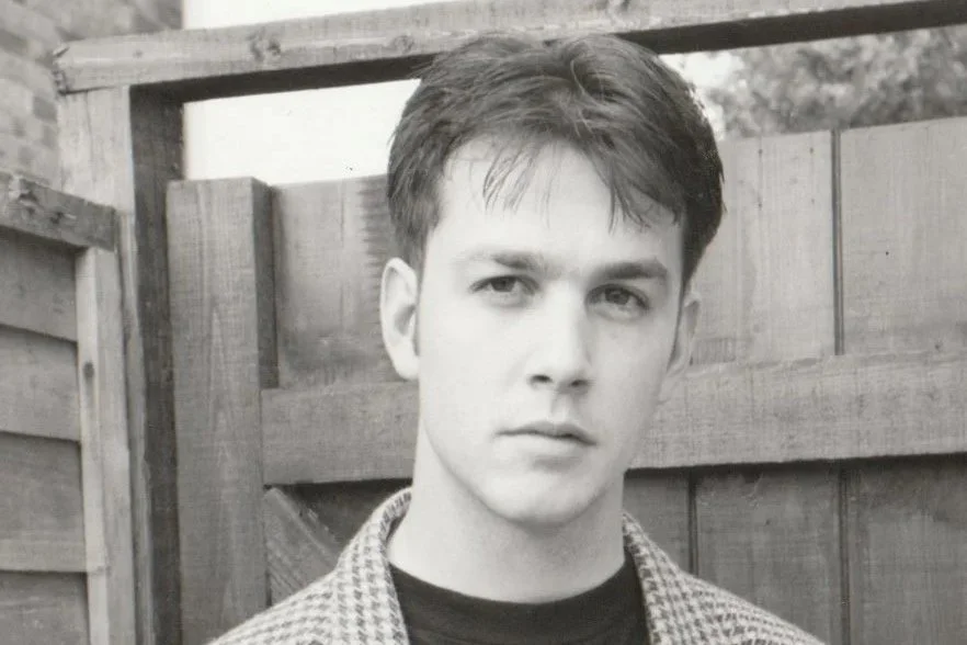 A young man with dark hair wearing a patterned jacket standing in front of a wooden fence.