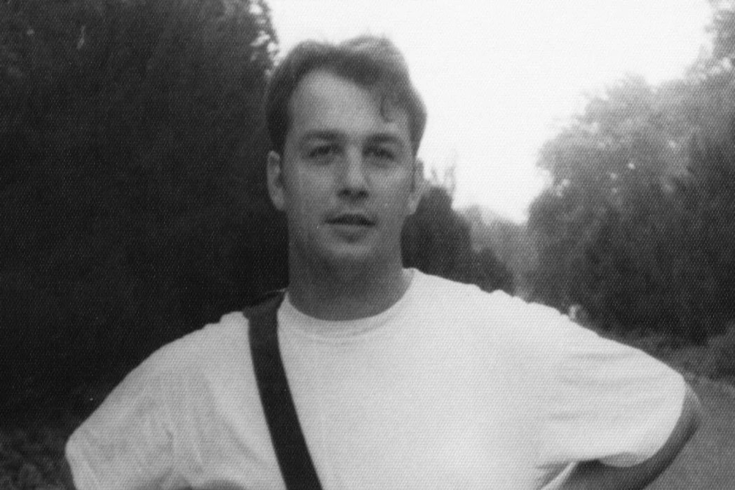 Black and white photo of a young man standing outdoors, wearing a white t-shirt and carrying a bag strap over his shoulder, with trees in the background.