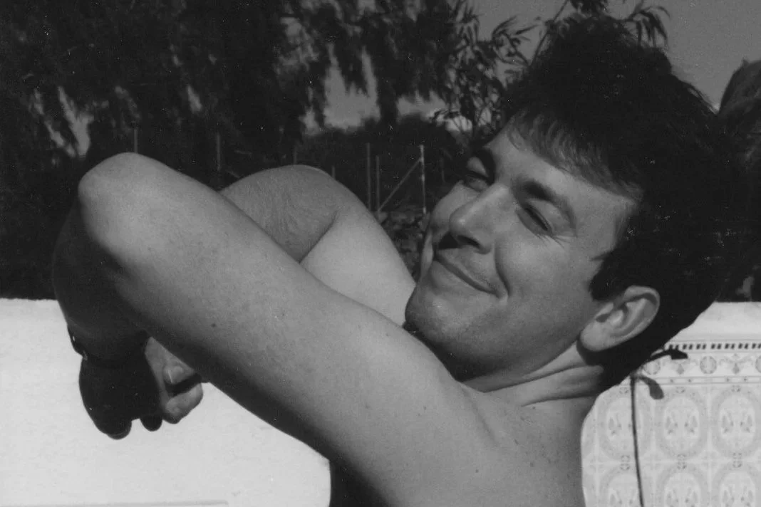 Young man with short dark hair smiling, flexing his arm, outdoors with trees in background, black and white photo.
