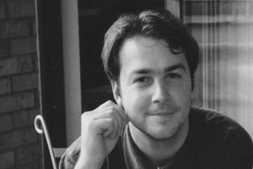 Black and white photo of a young man with dark hair, smiling softly, resting his head on his hand, in front of a brick wall in an indoor setting.