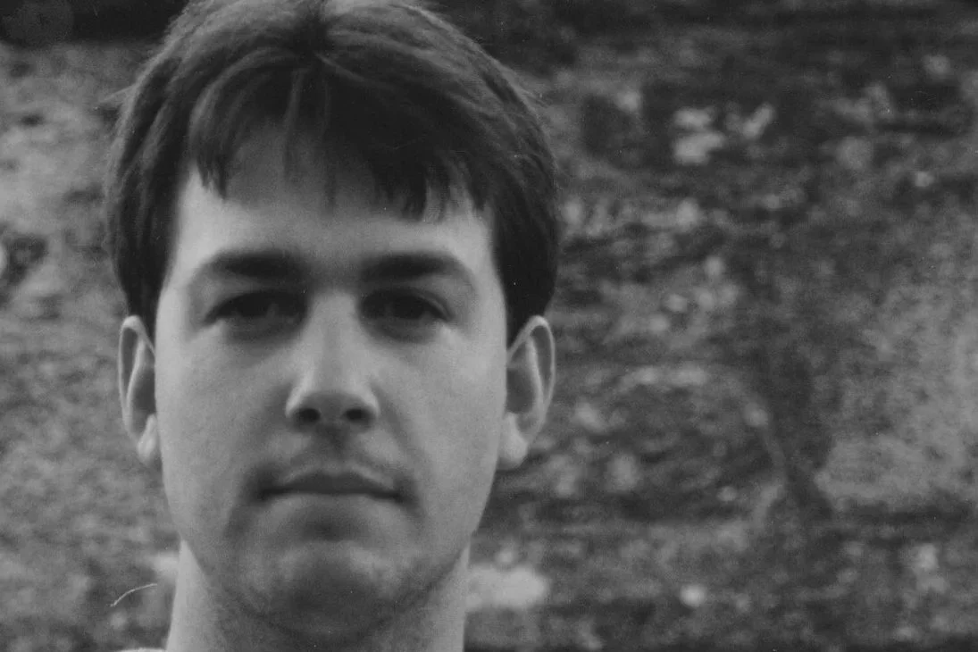 Black and white close-up photo of a young man with dark hair, looking directly at the camera, with a blurred natural background.