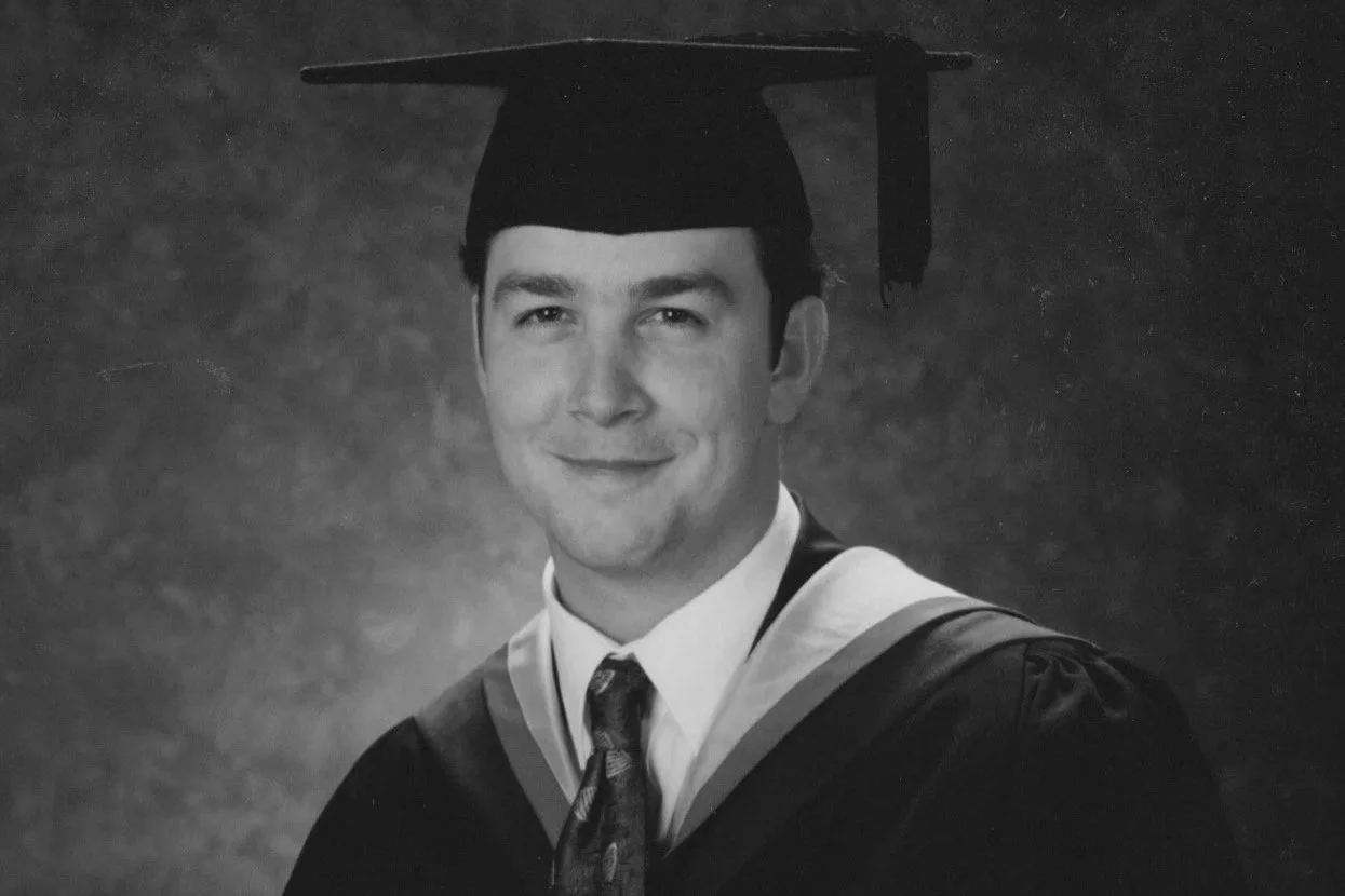 Black and white portrait of a young man in academic cap and gown smiling, with a background gradient.