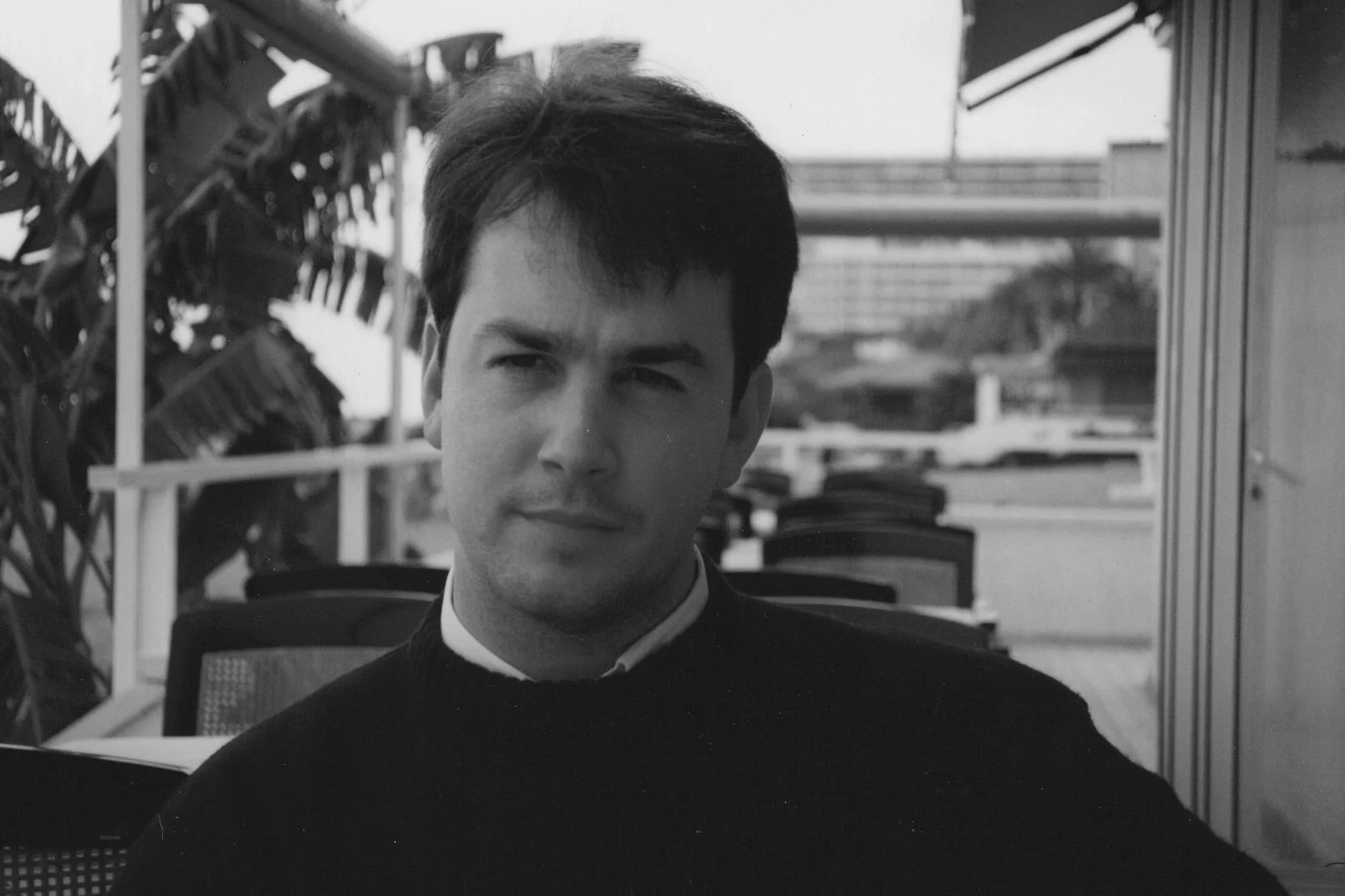 Black and white photo of a man with short dark hair, wearing a dark sweater over a collared shirt, sitting outdoors on a patio with patio furniture and potted plants.