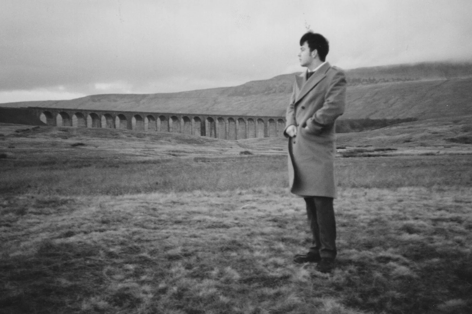 A young man standing in an open field with a long coat, hands in pockets, looking to the side, with a stone arch bridge and hills in the background.