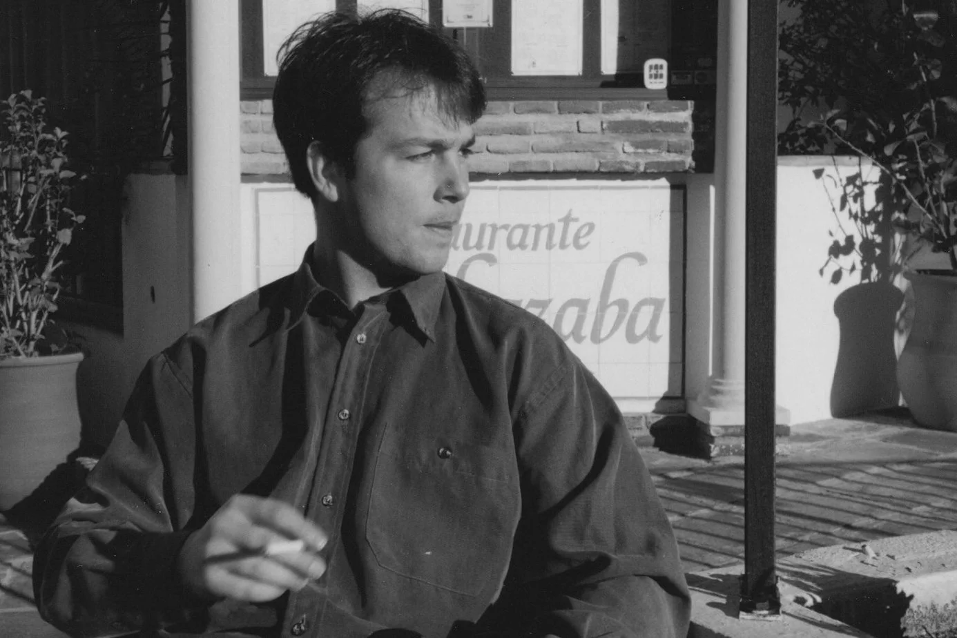 A young man with dark hair, wearing a button-up shirt, sitting outside a restaurant with a sign in the background that reads "Restaurante Chaba," holding a cigarette.
