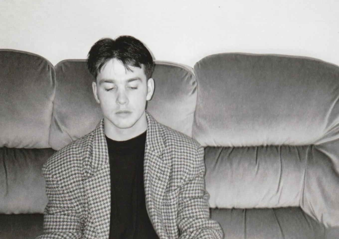 Young man with dark hair and closed eyes sitting on a cushioned sofa with a checkered blazer and a black shirt.