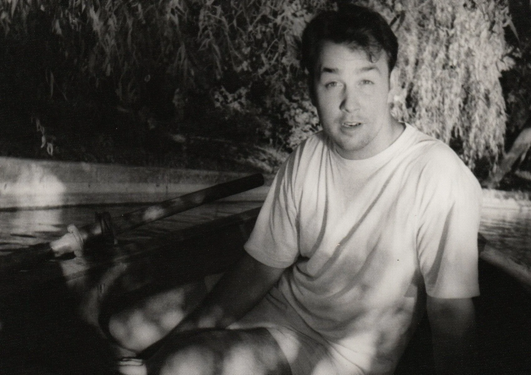 Black and white photo of a young man sitting on a boat near a rocky shoreline, looking at the camera with a relaxed expression, with trees and bushes in the background.