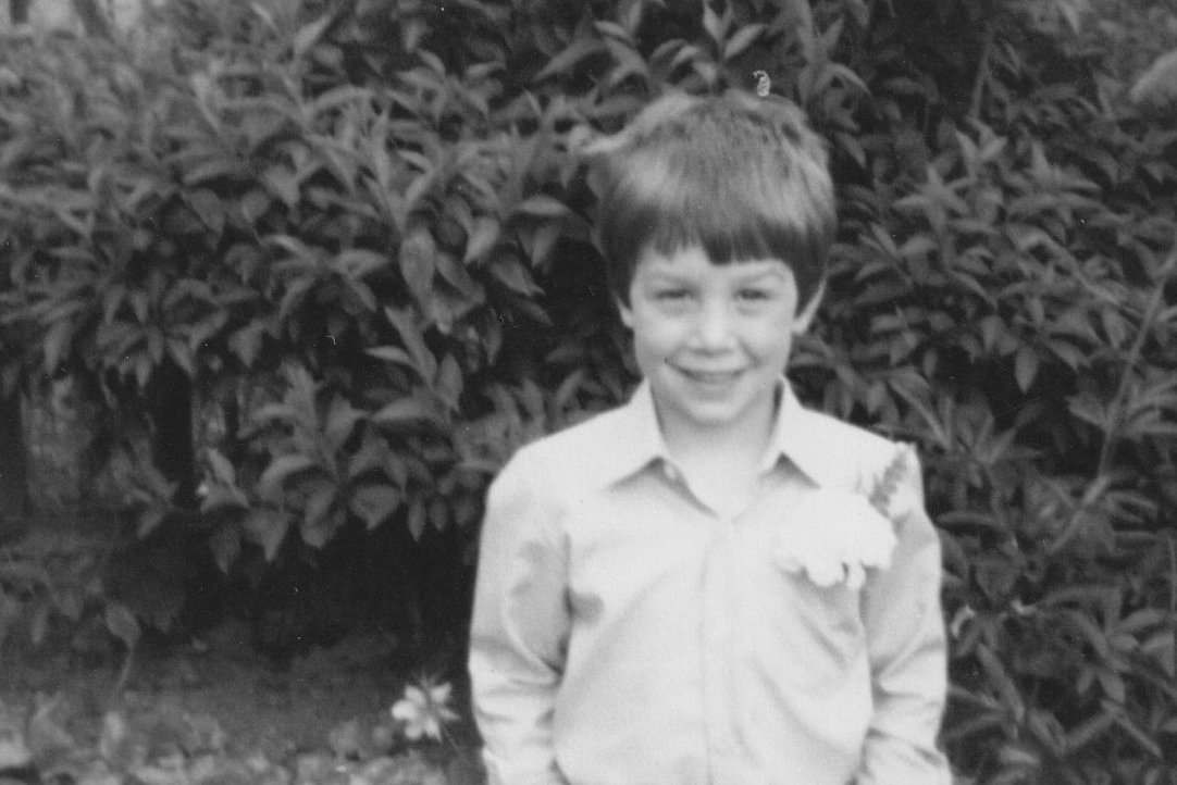 A young boy standing outdoors in front of a bush, smiling at the camera, wearing a light-colored shirt.