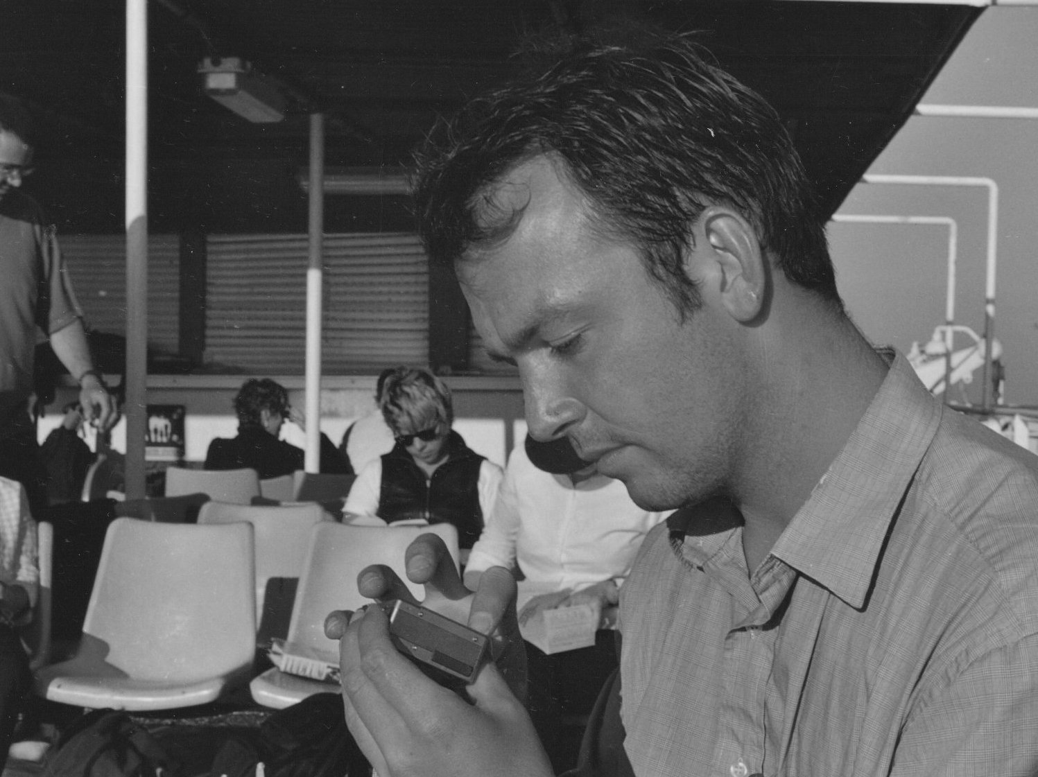 A man with dark, curly hair looking down at his phone on a sunny day at an outdoor seating area of an airport or bus terminal, with several people sitting and standing in background.
