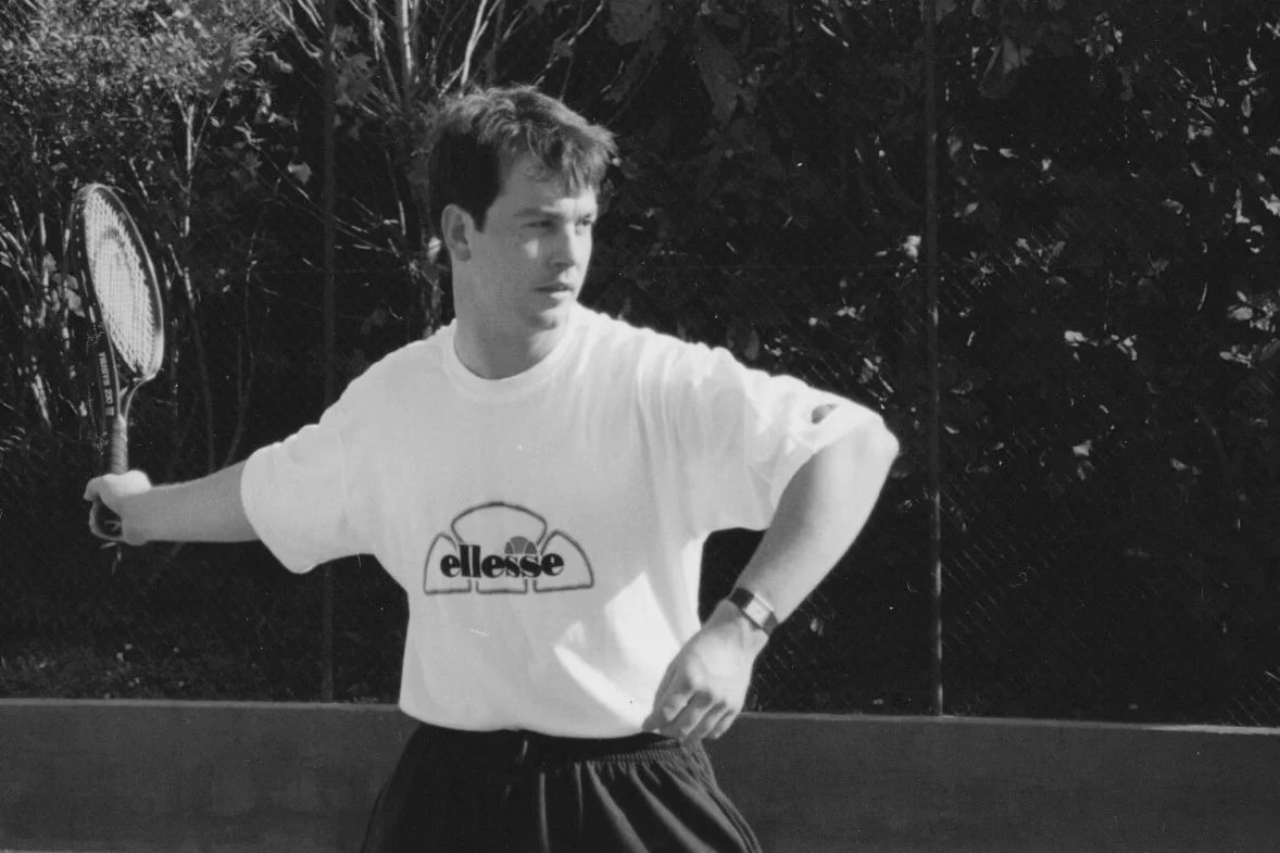 A young man is playing tennis outdoors, wearing a white t-shirt with the 'Ellesse' logo, holding a tennis racket, and preparing to serve or hit the ball, with a fence and foliage in the background.