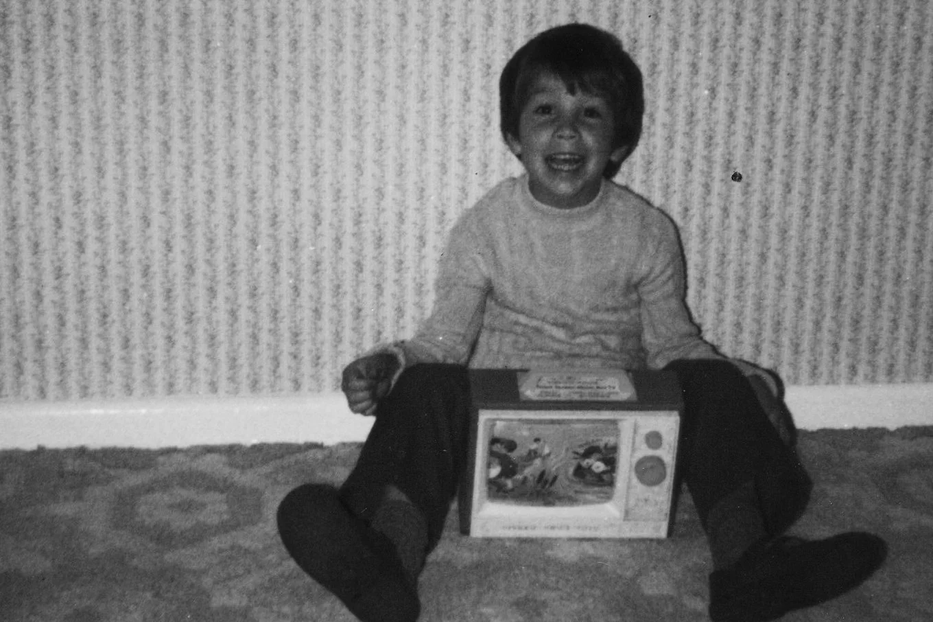 A young boy with dark hair, sitting on a carpeted floor, smiling, holding a toy box with cartoon characters on it.