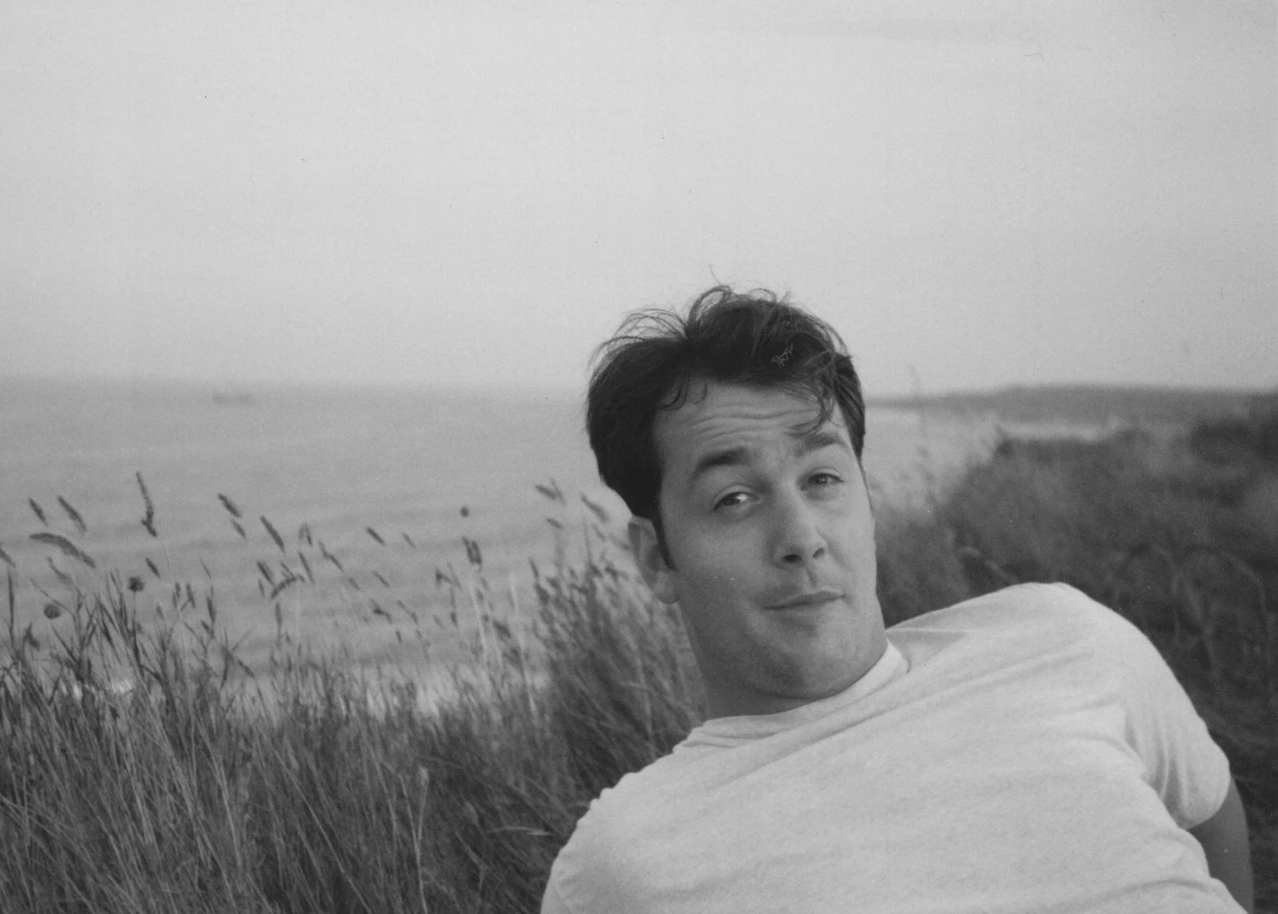 A young man with dark hair wearing a light-colored shirt, lying on his side outdoors near a grassy area by the water, looking at the camera with a slight smile, black and white photograph.
