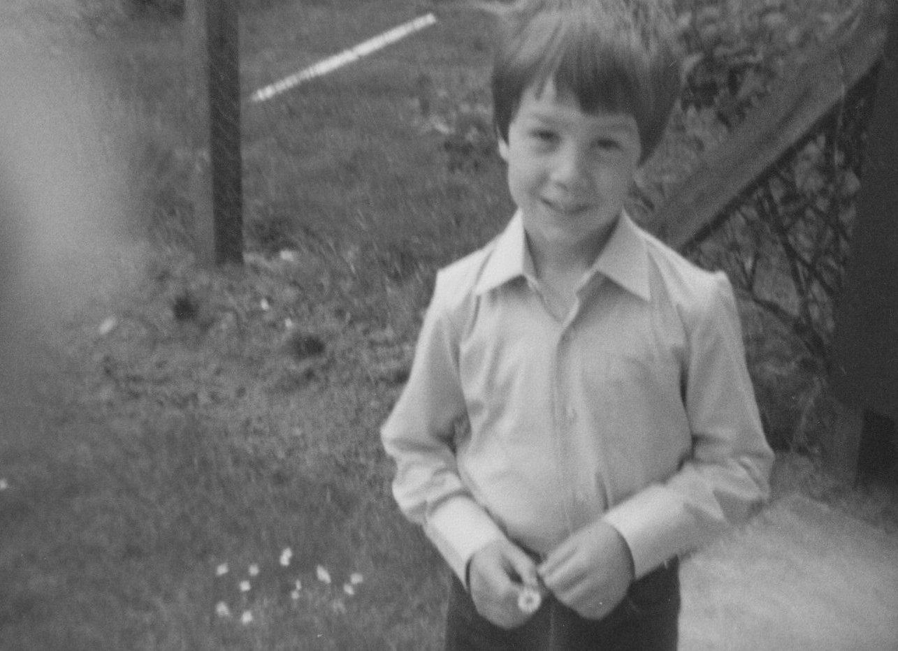 A young boy with short dark hair, wearing a long-sleeved button-up shirt and dark pants, standing outdoors near a staircase and a fence.